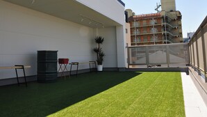 Um terraço no telhado com grama sintética, uma área de estar com cadeiras, uma lixeira e um vaso de planta.