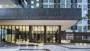 Un bâtiment moderne doté de grandes fenêtres en verre, d’un hall d’entrée spacieux et d’un espace extérieur aménagé avec des sièges, le tout baigné de lumière.