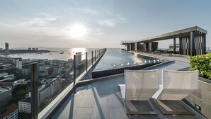 Une piscine à débordement située sur le toit, offrant une vue sur la ville, ainsi qu’un bâtiment à l’architecture moderne et des chaises longues.