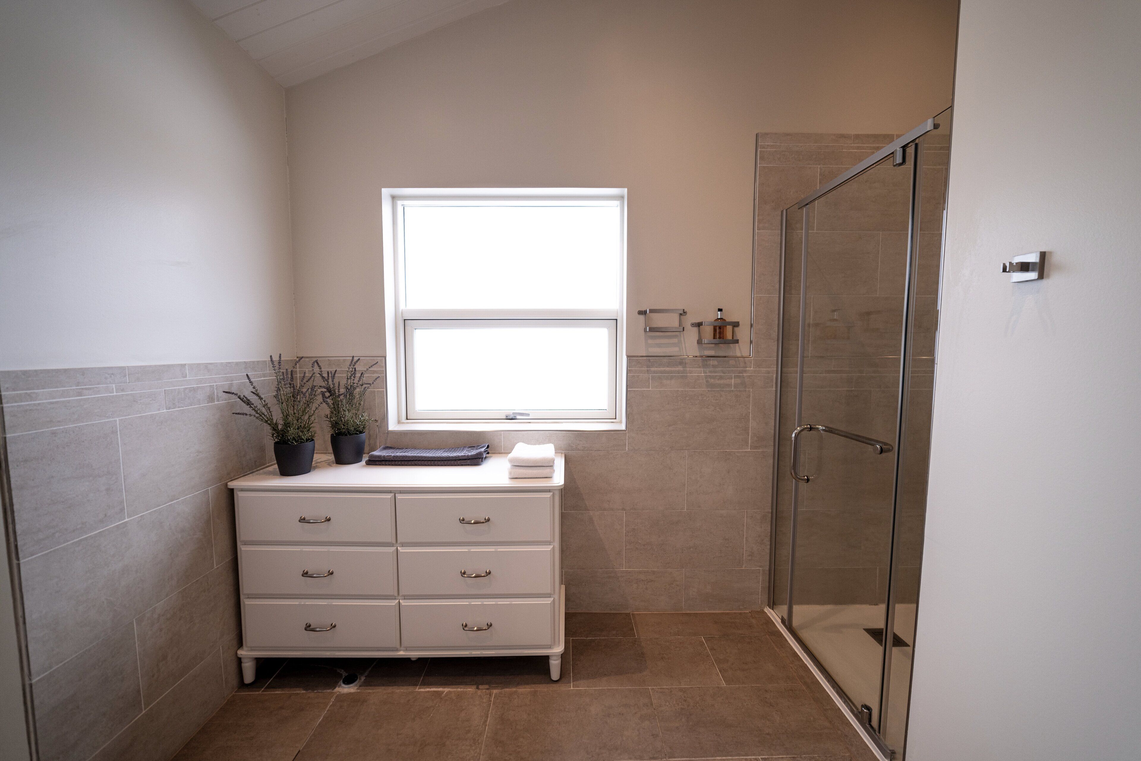 A bathroom with a white vanity, a window, a glass shower enclosure, and a tiled floor.