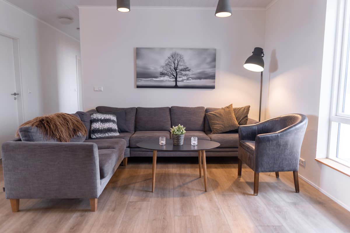 Ein modernes Wohnzimmer mit einem grauen Ecksofa, einem runden Couchtisch und einem grauen Sessel. Eine Stehlampe und ein Baumgemälde sind an der Wand zu sehen.
