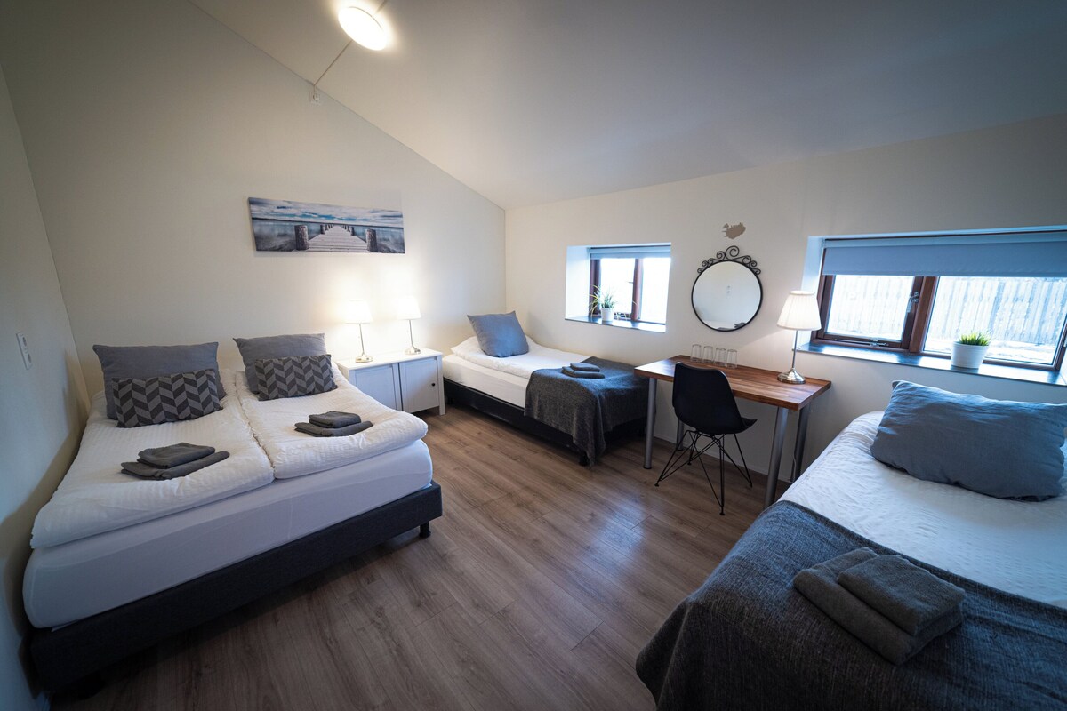 A modern bedroom with two beds, a desk, and a chair. There are windows providing natural light and a view of the outside.
