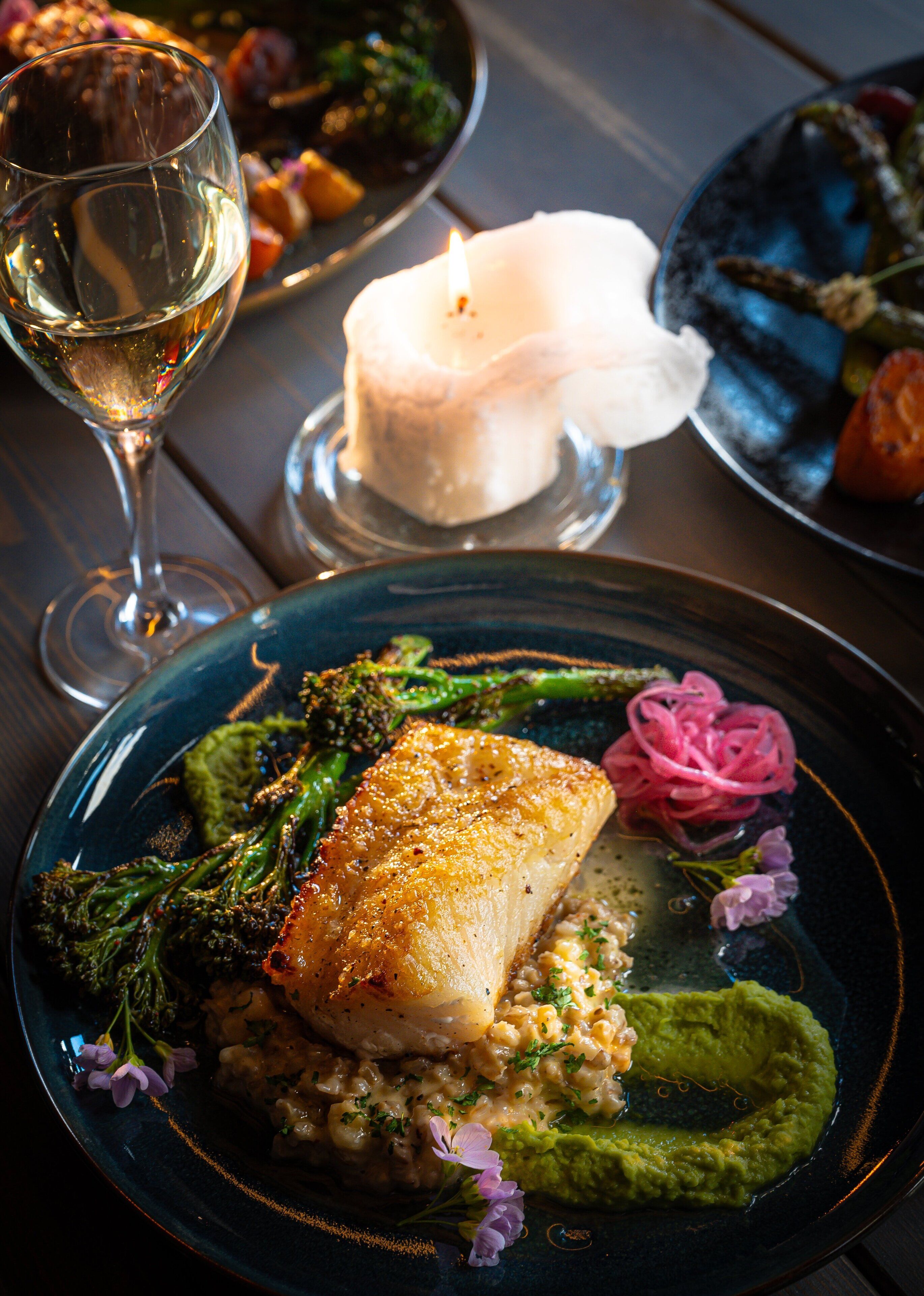 A plate of food with a fish fillet, broccoli, and a creamy sauce, accompanied by a glass of wine and a lit candle.