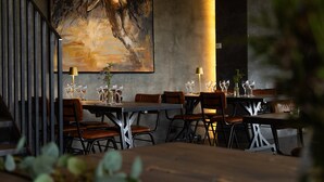 A restaurant with wooden tables and chairs, a large painting on the wall, and a staircase.