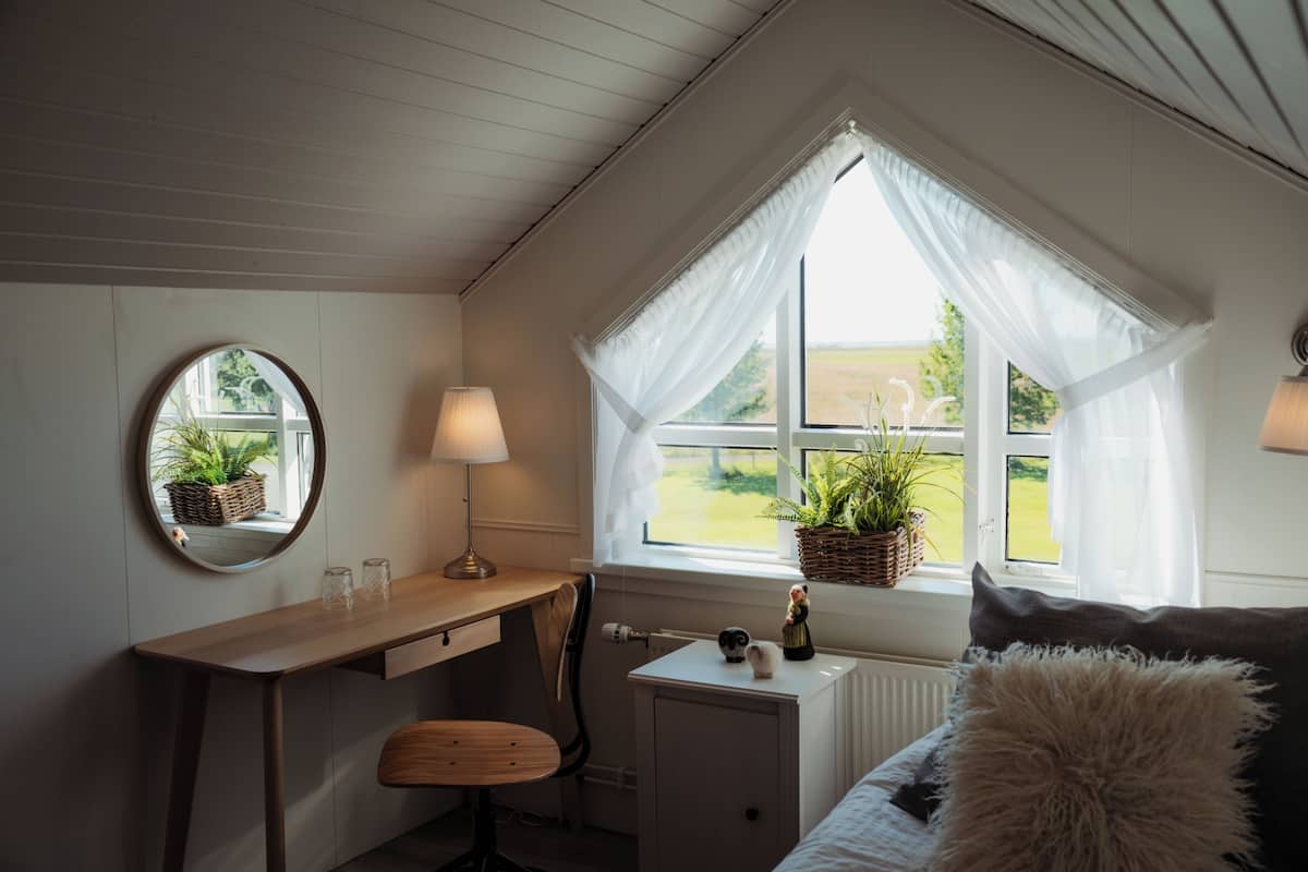 Ein gemütliches Schlafzimmer mit einem Holztisch, einem runden Spiegel, einem kleinen Stuhl, einem Nachttisch mit Lampe und einem Fenster mit Blick ins Grüne.