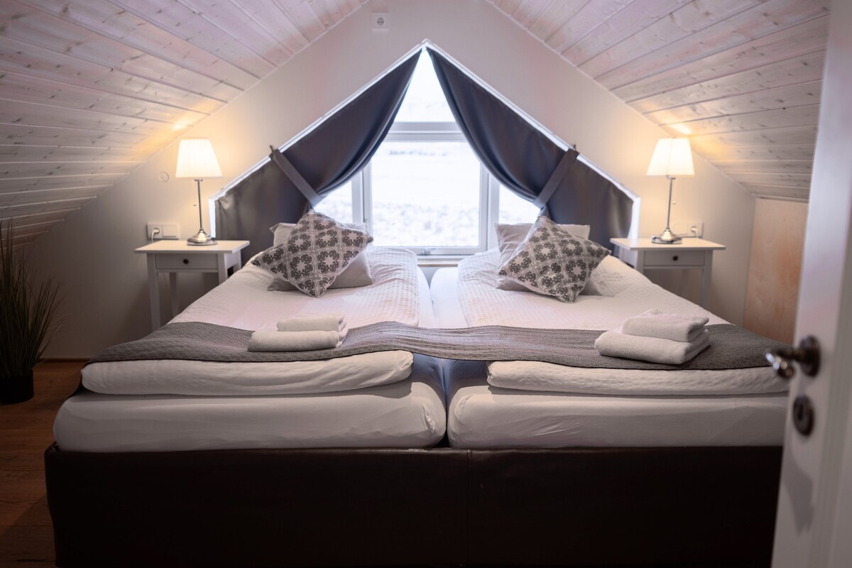 A bedroom with a loft-style ceiling, two bedside tables with lamps, a bed with a duvet and pillows, and a window with curtains.