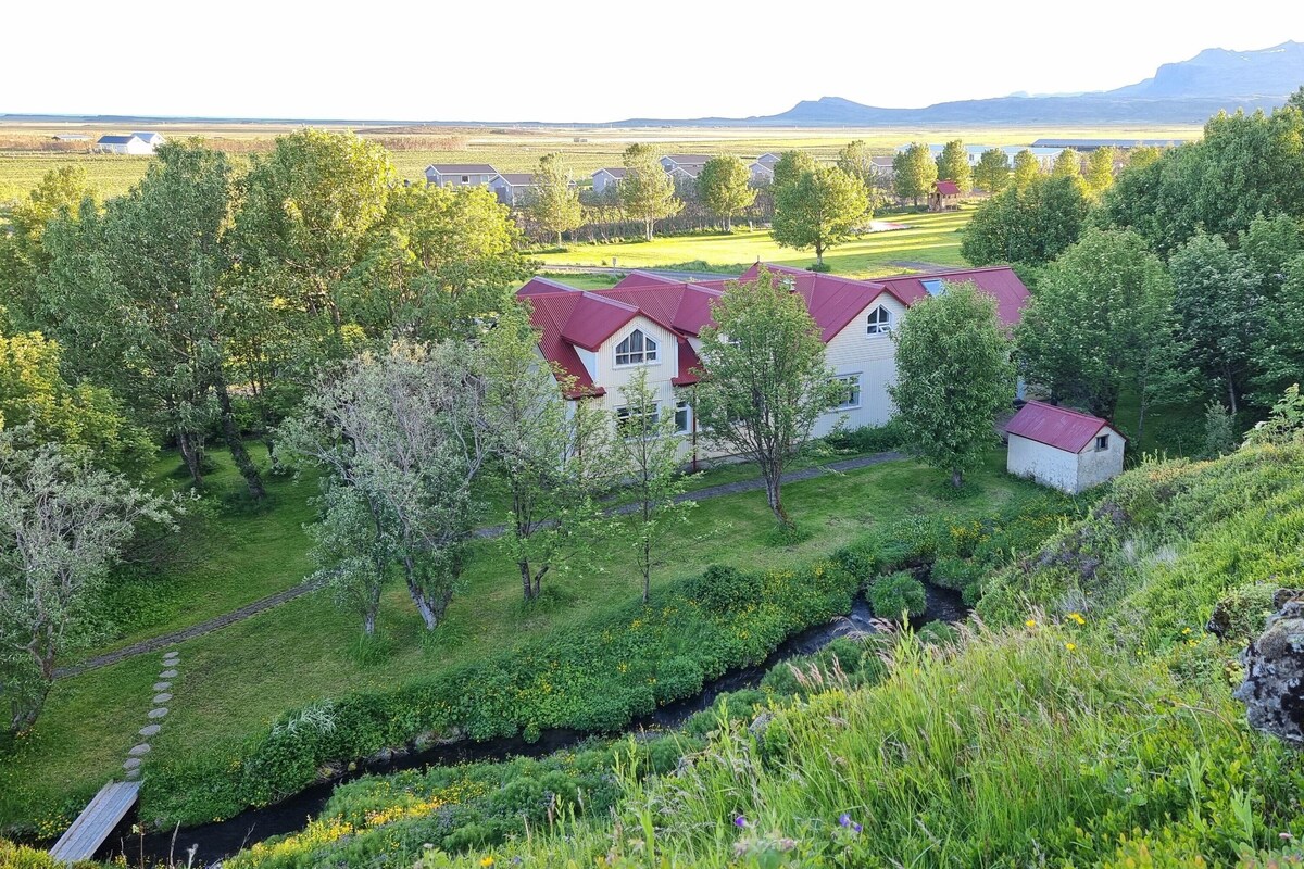 A rural landscape with a house surrounded by lush greenery and a small stream.