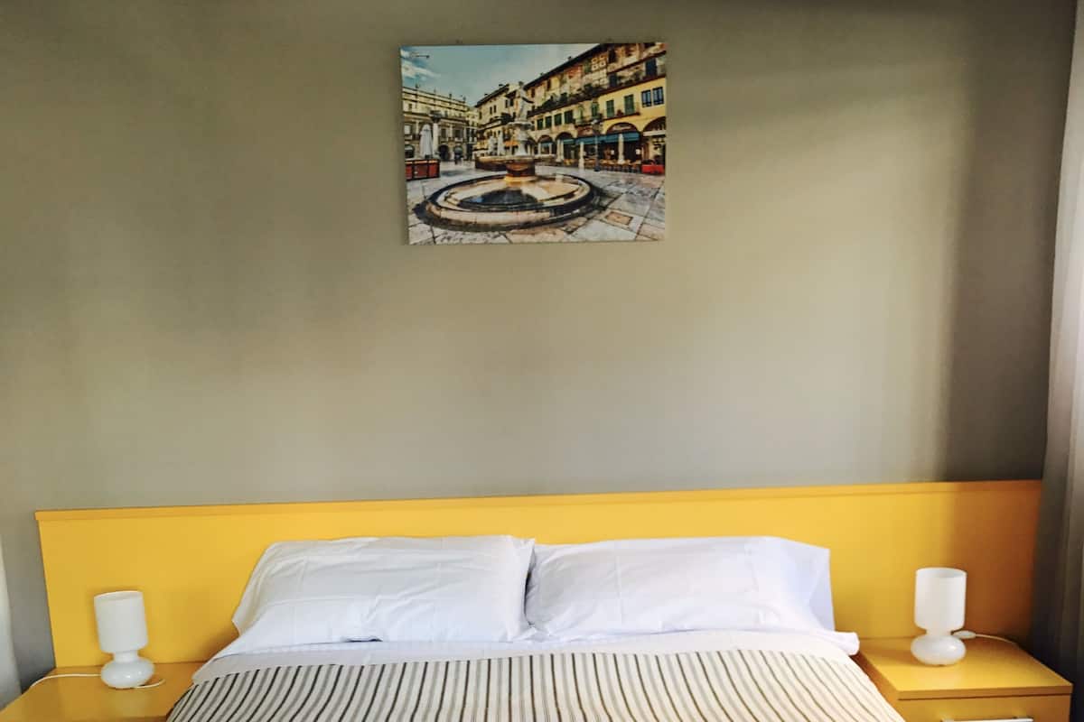 A hotel room with a bed, two bedside tables, a nightstand, a painting on the wall, and a window.
