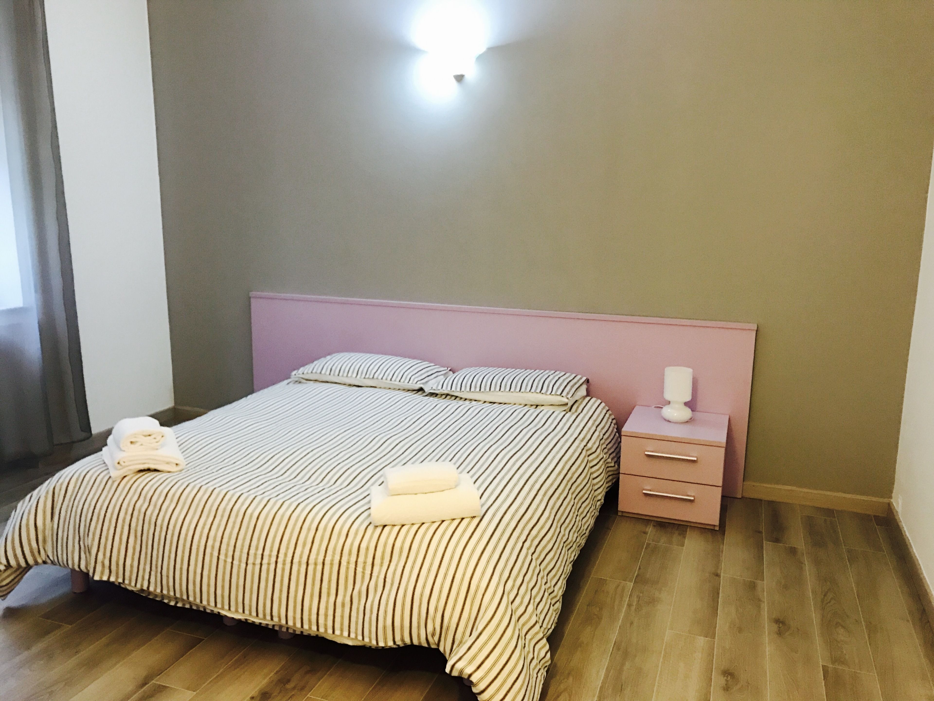 A neatly made bed with striped bedding, a pink headboard, and a bedside table with a lamp.