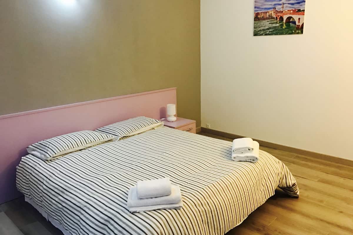 A neatly made bed with striped bedding, a pink headboard, and a small bedside table with a lamp and a folded towel.