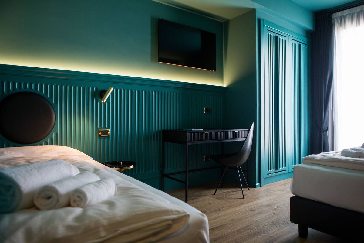 Una habitación de hotel con cama, escritorio, silla, televisor y ventana con cortinas.