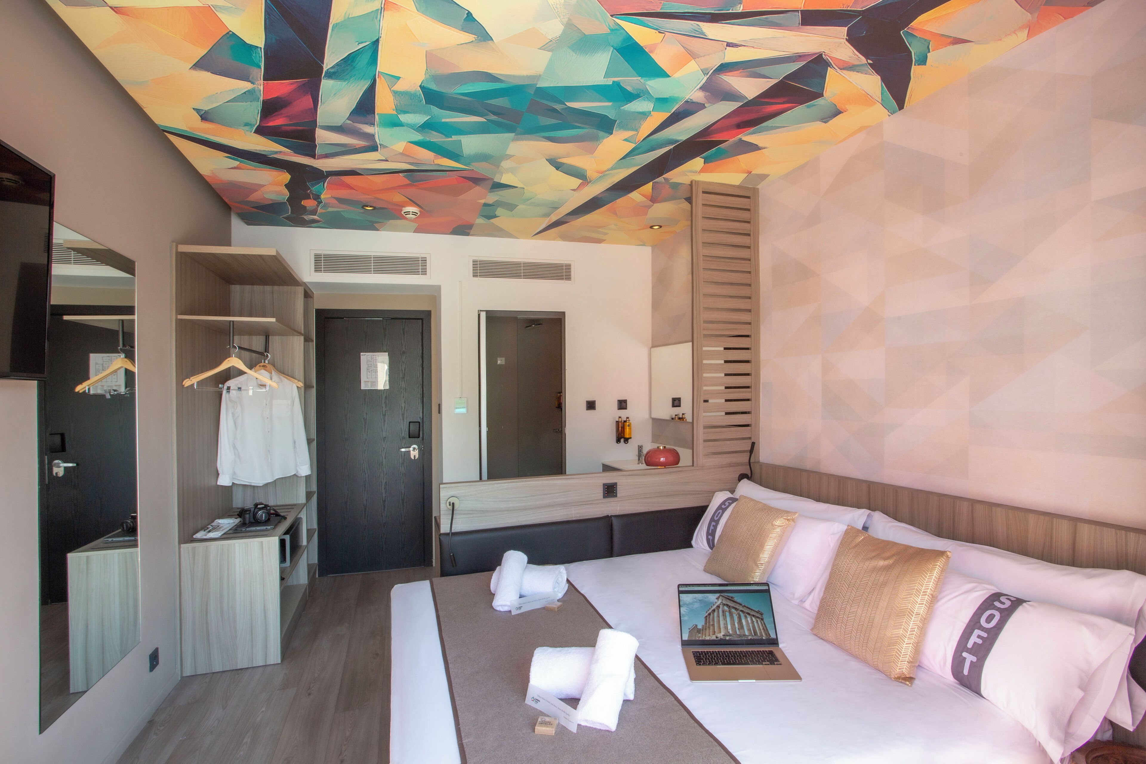 A modern hotel room with a geometric patterned ceiling, a large bed with pillows, a laptop, and a bedside table with a red object.