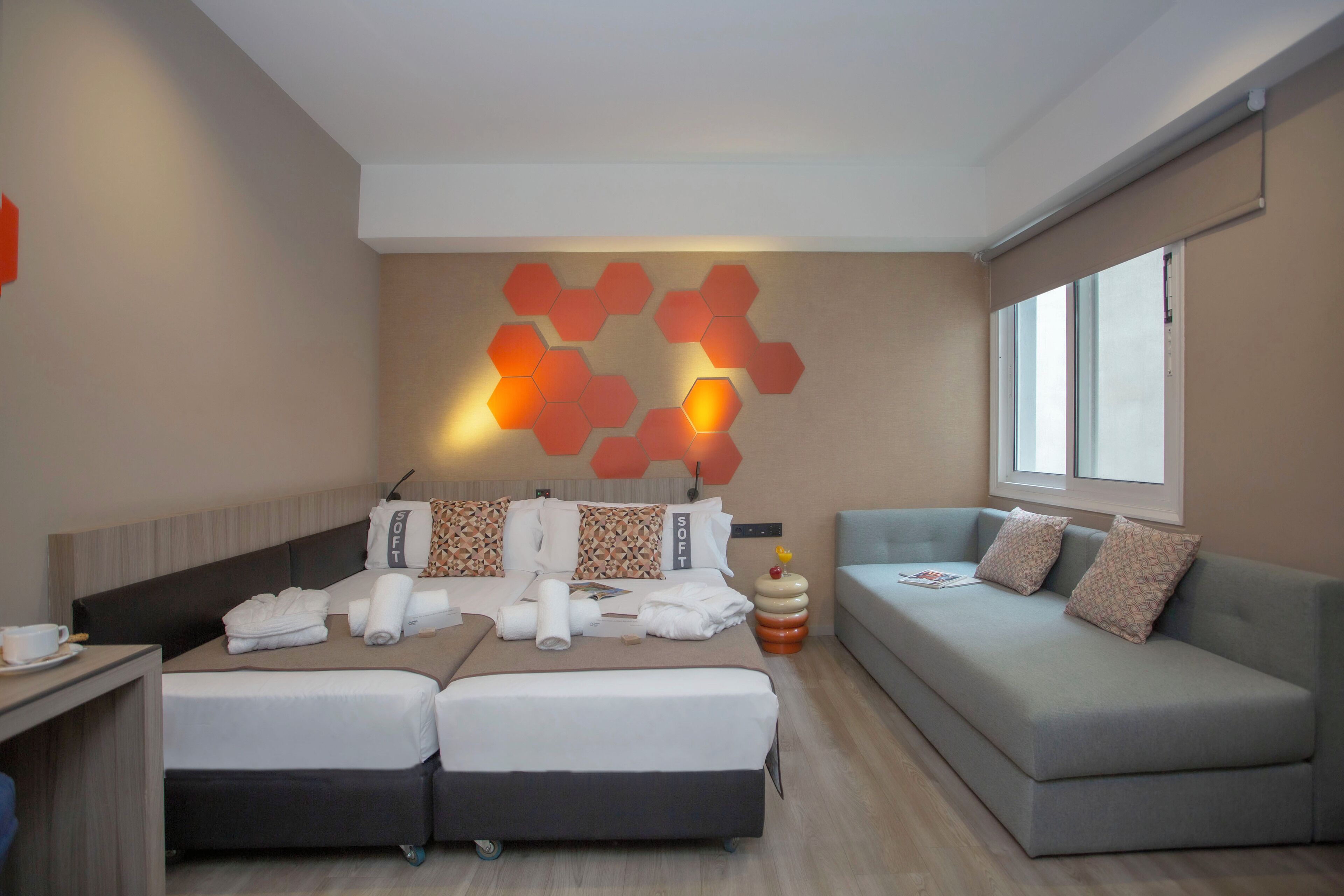 A modern hotel room with a bed, a sofa, a desk, and a wall with hexagonal light panels.