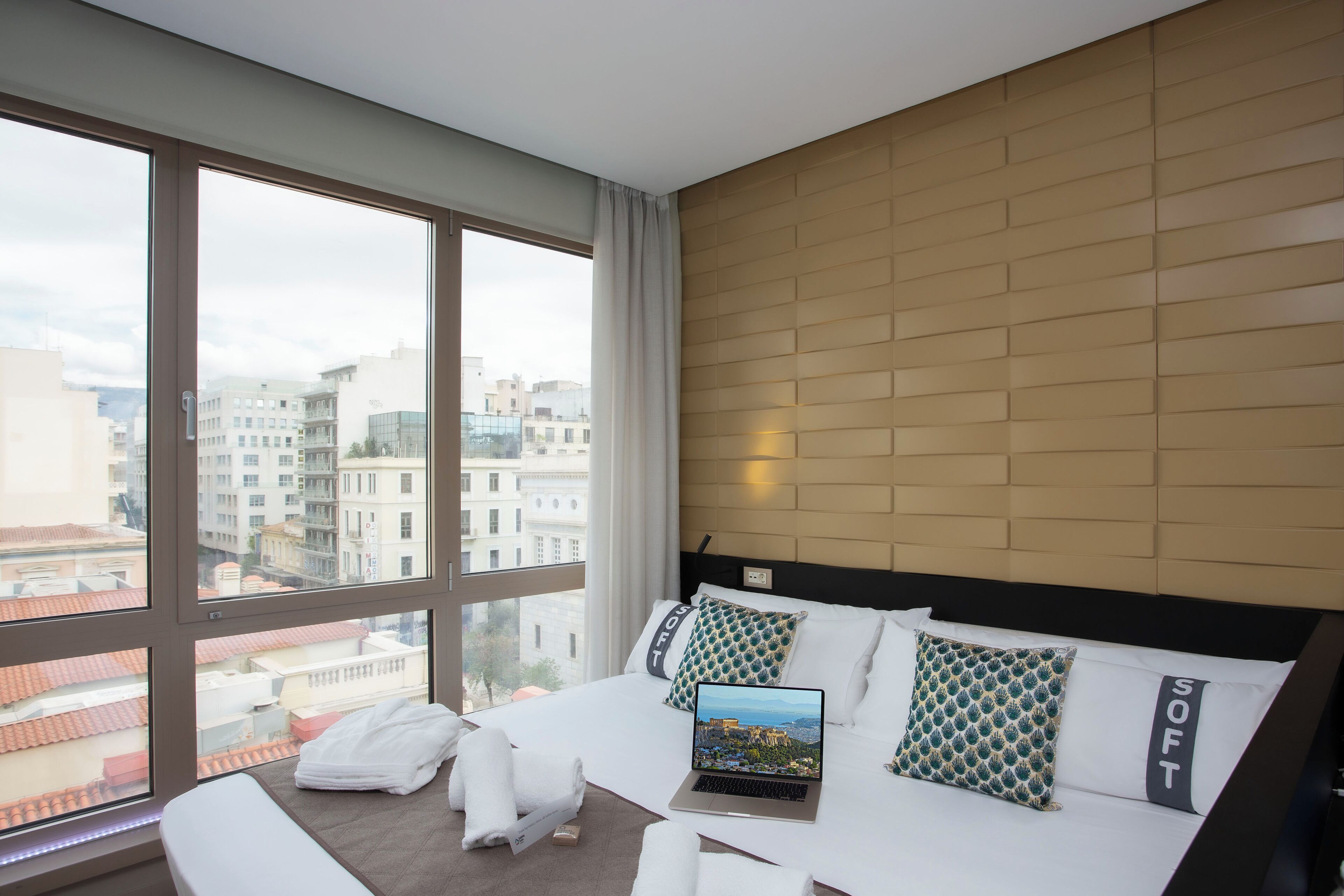 A modern hotel room with a large window, a bed with white linens, a laptop, and a city view.