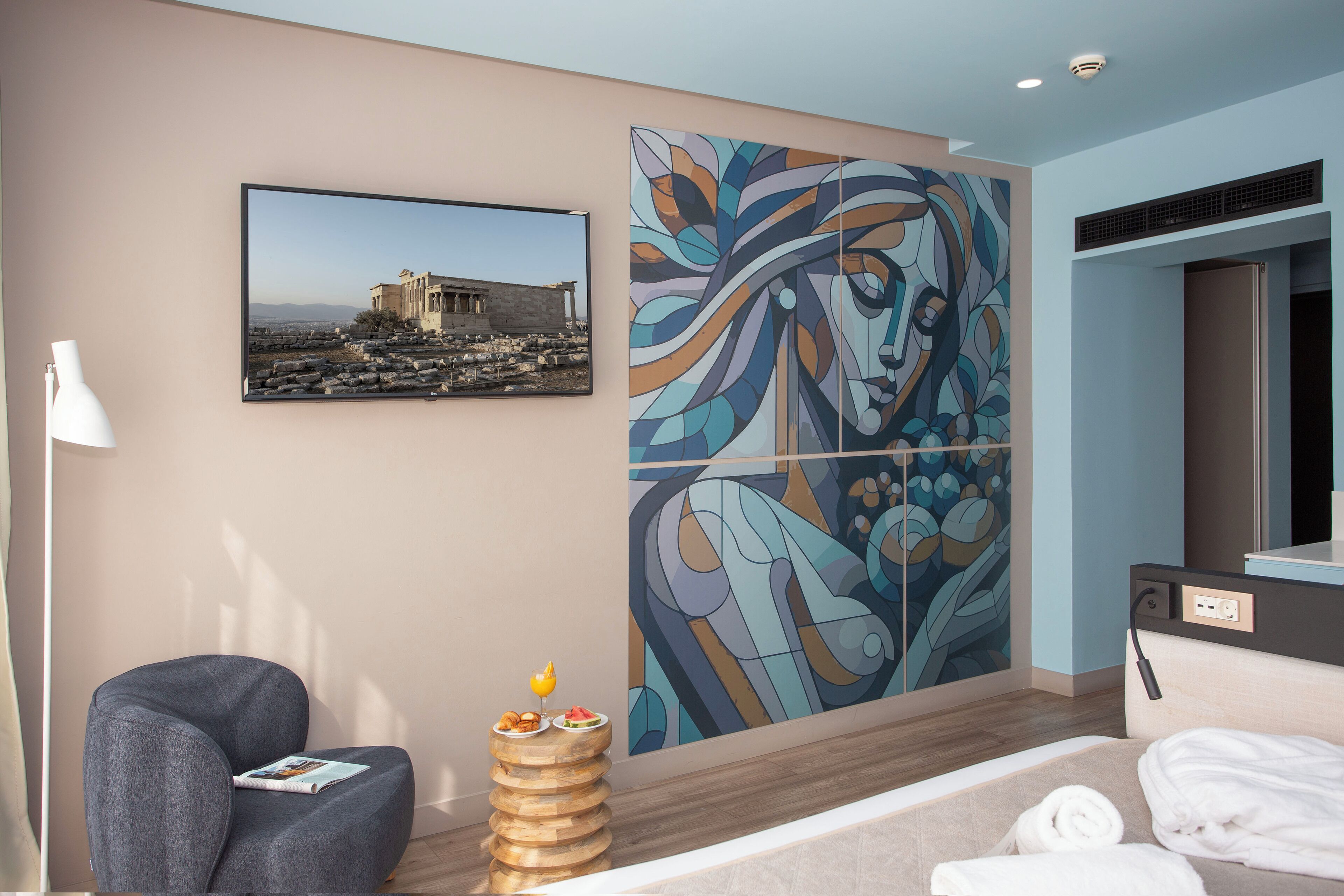 Una habitación de hotel moderna con un cuadro abstracto grande, una fotografía enmarcada de una estructura antigua, un sillón cómodo, una pequeña mesa auxiliar de madera con un frutero y toallas dobladas ordenadamente sobre la cama.