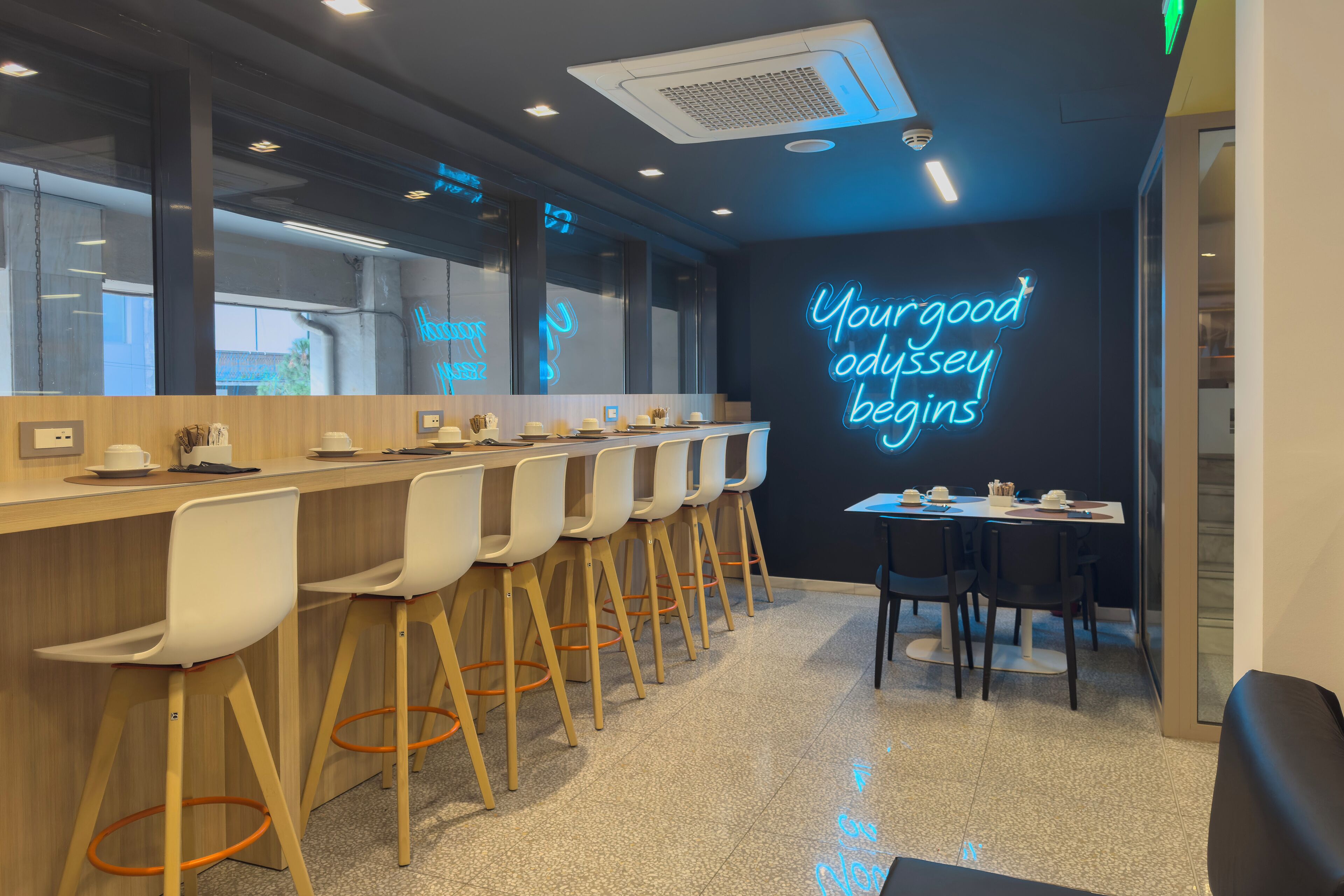 Un área de bar moderna con un letrero de neón que dice "Your good odyssey begins", taburetes de bar y una mesa con sillas.