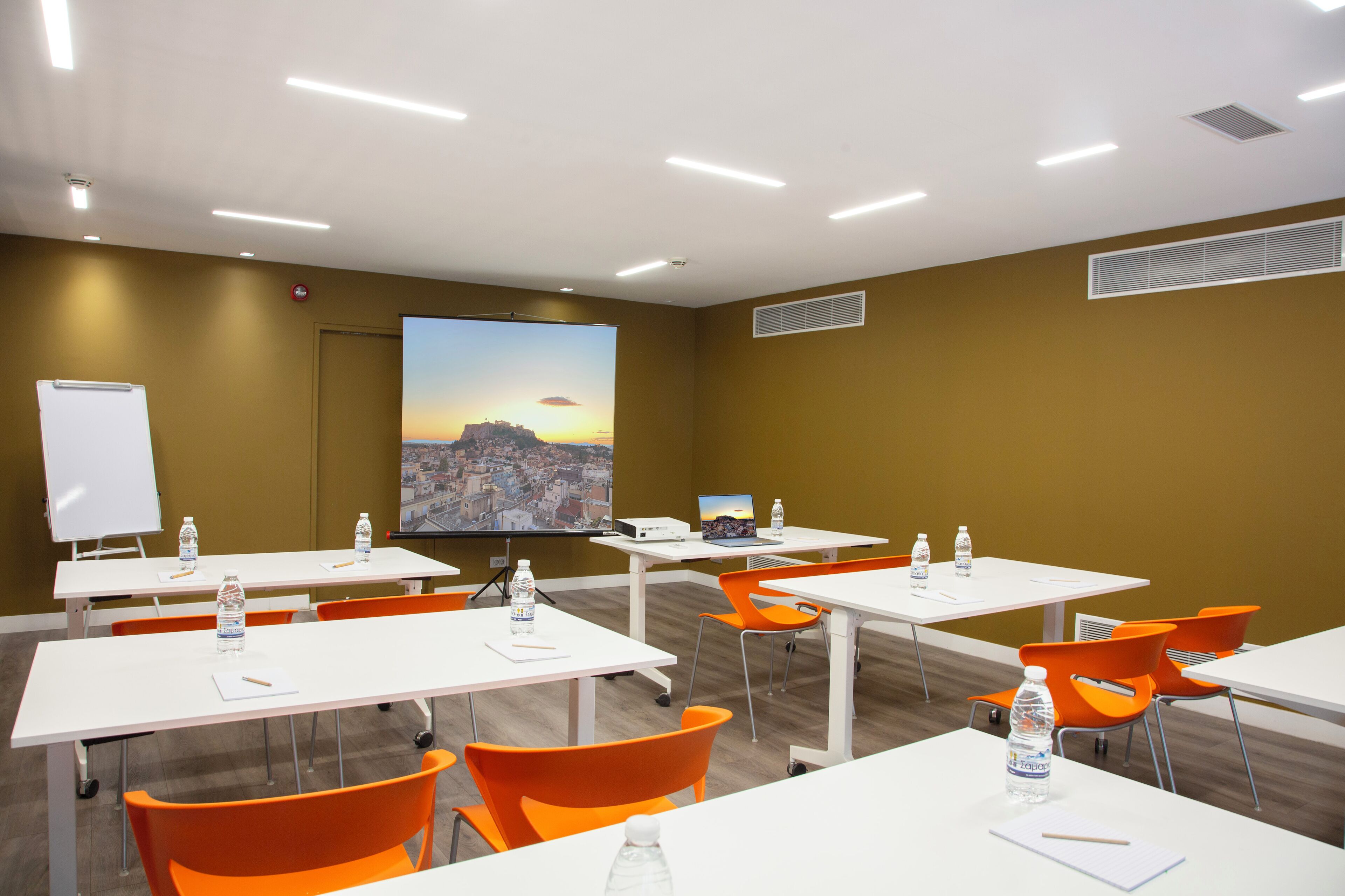 A conference room with white tables, orange chairs, and a large screen displaying a sunset cityscape.