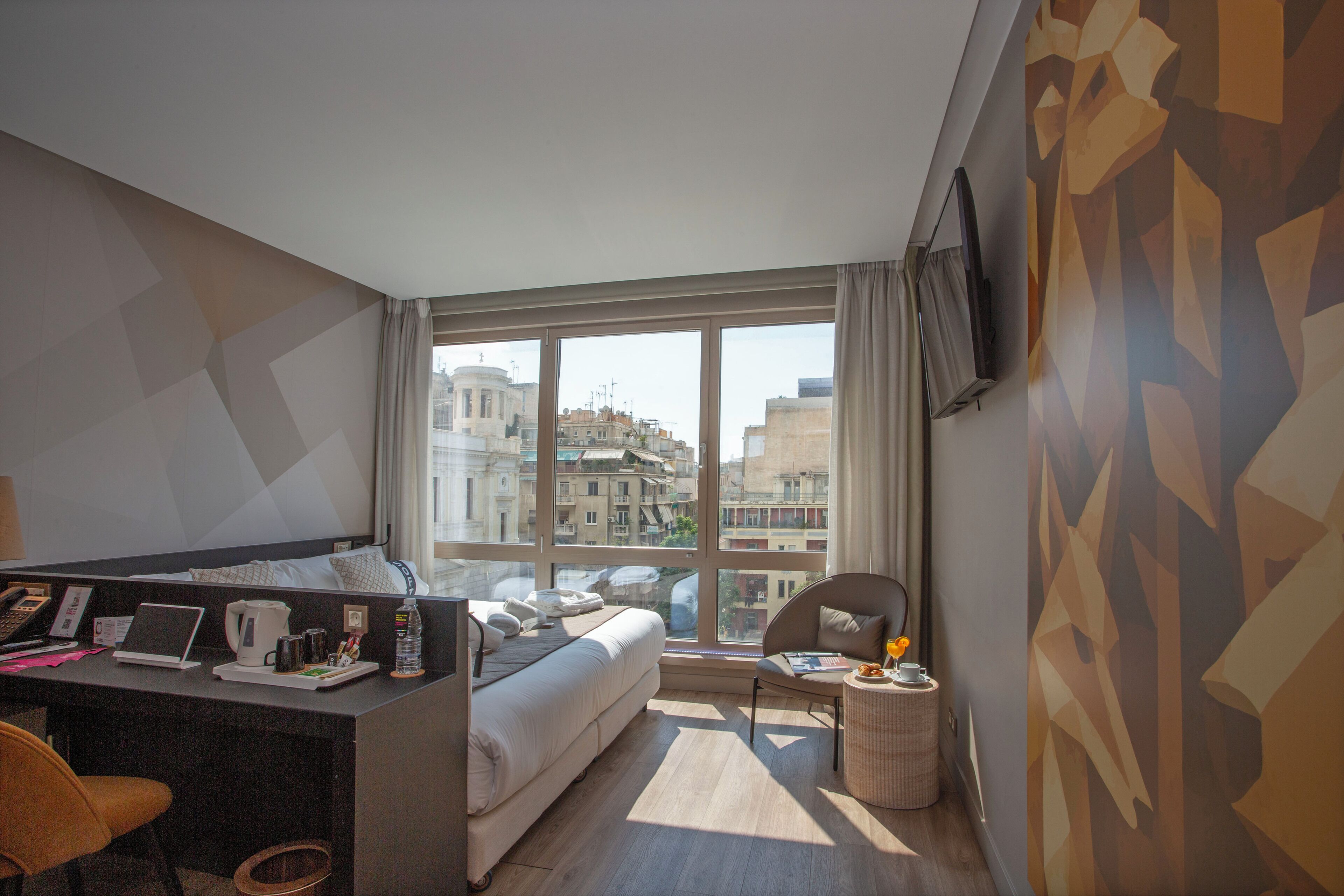 A modern hotel room with a large window, a desk with a laptop, a bed, and a small round table with a chair.