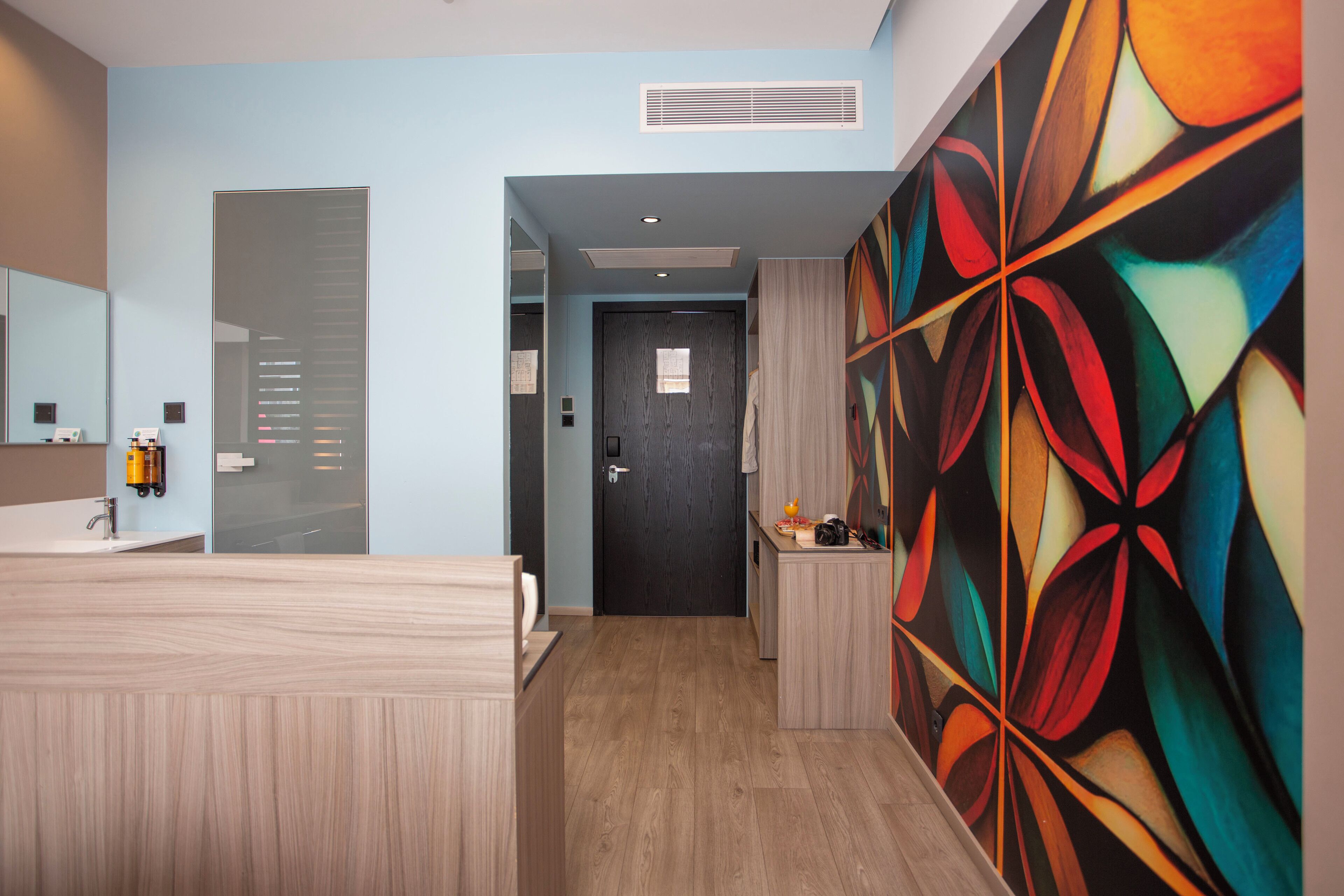 Un baño de hotel moderno con un tocador de madera, un espejo grande y un diseño abstracto y colorido en la pared.