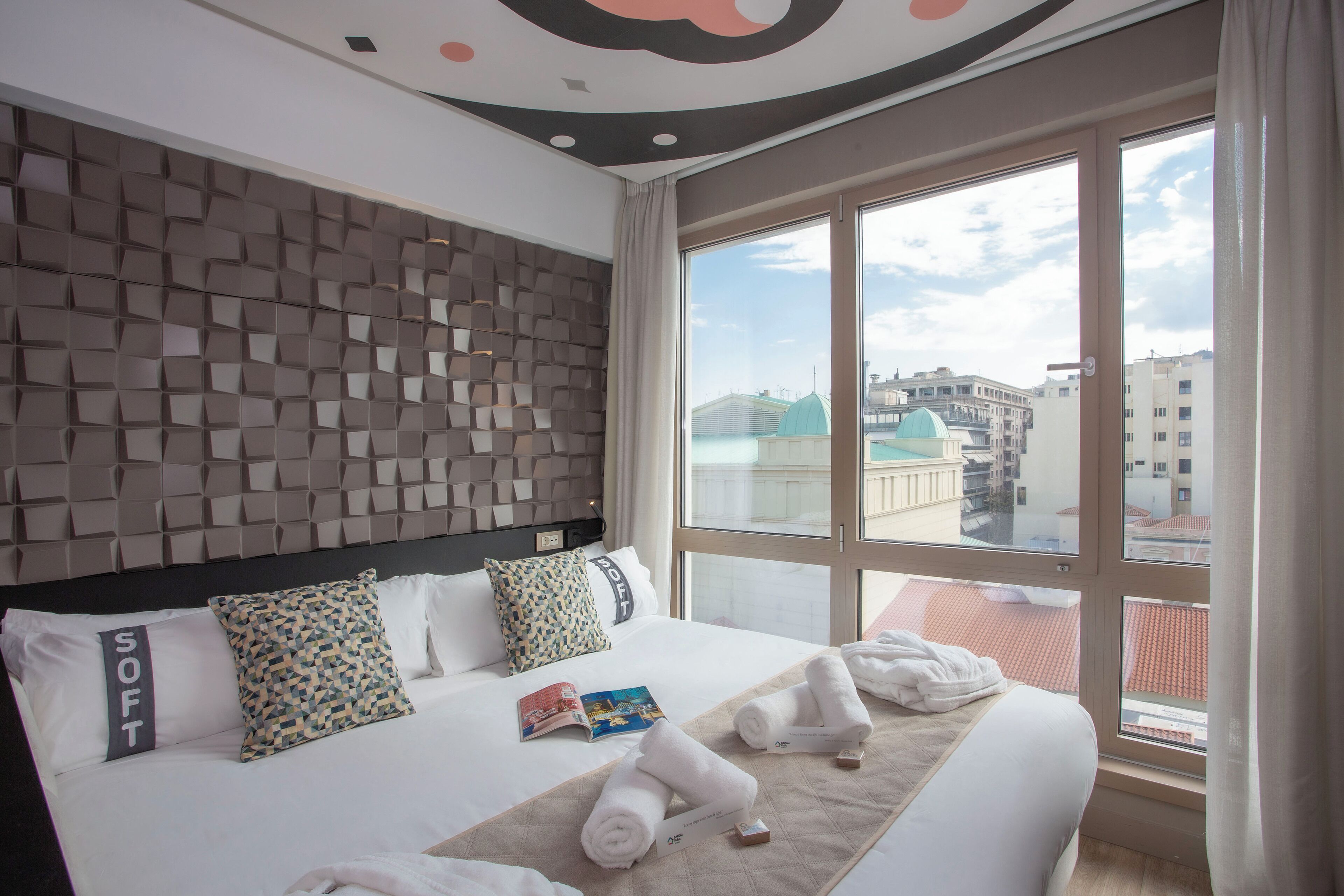 A hotel room with a large bed, multiple pillows, a quilt, and a view of the city through large windows.