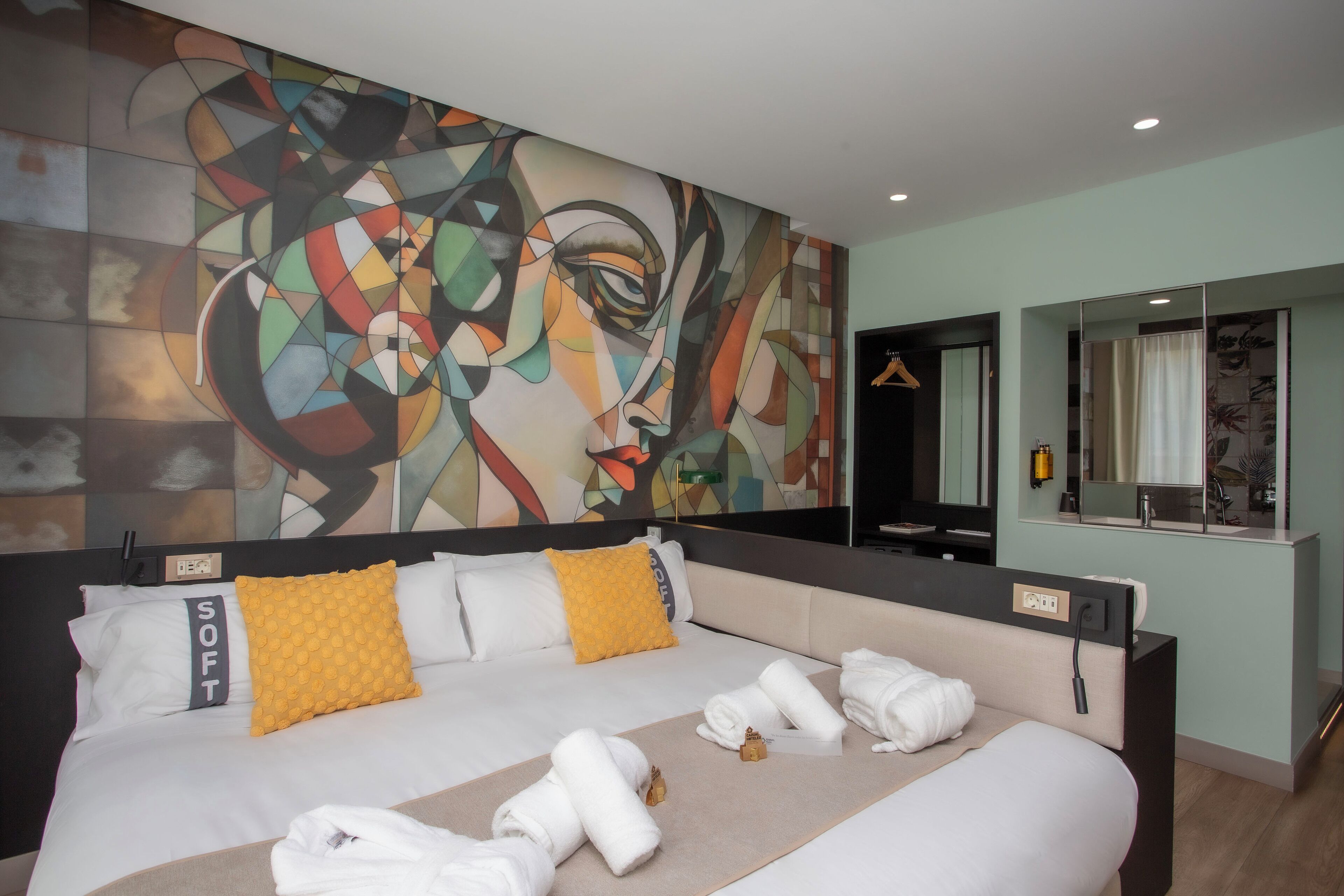 A modern hotel room with a large abstract painting on the wall, a bed with white linens and a yellow pillow, and a mirror above the bed.