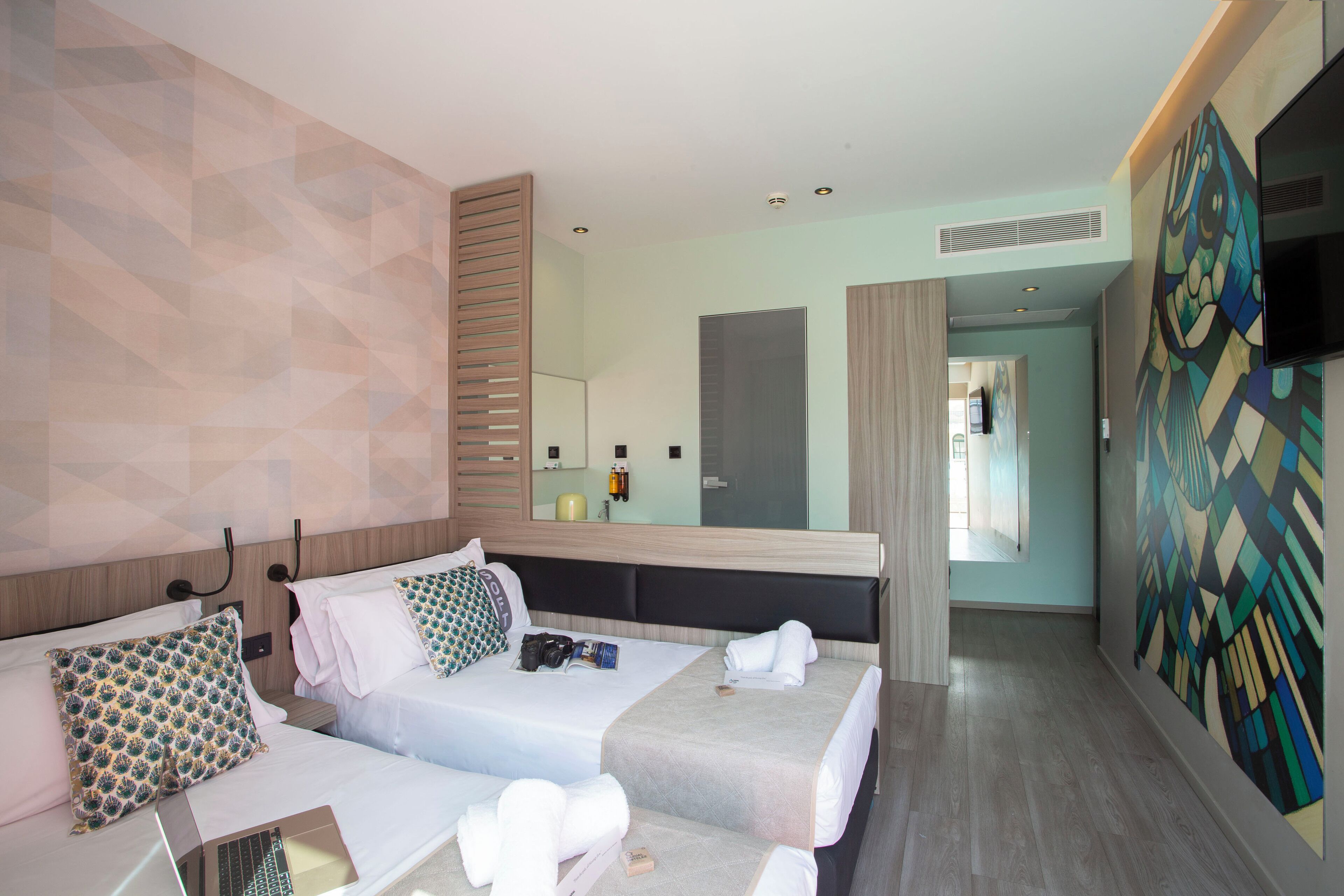 Habitación de hotel con dos camas, un espejo grande, un televisor y un diseño geométrico en la pared.