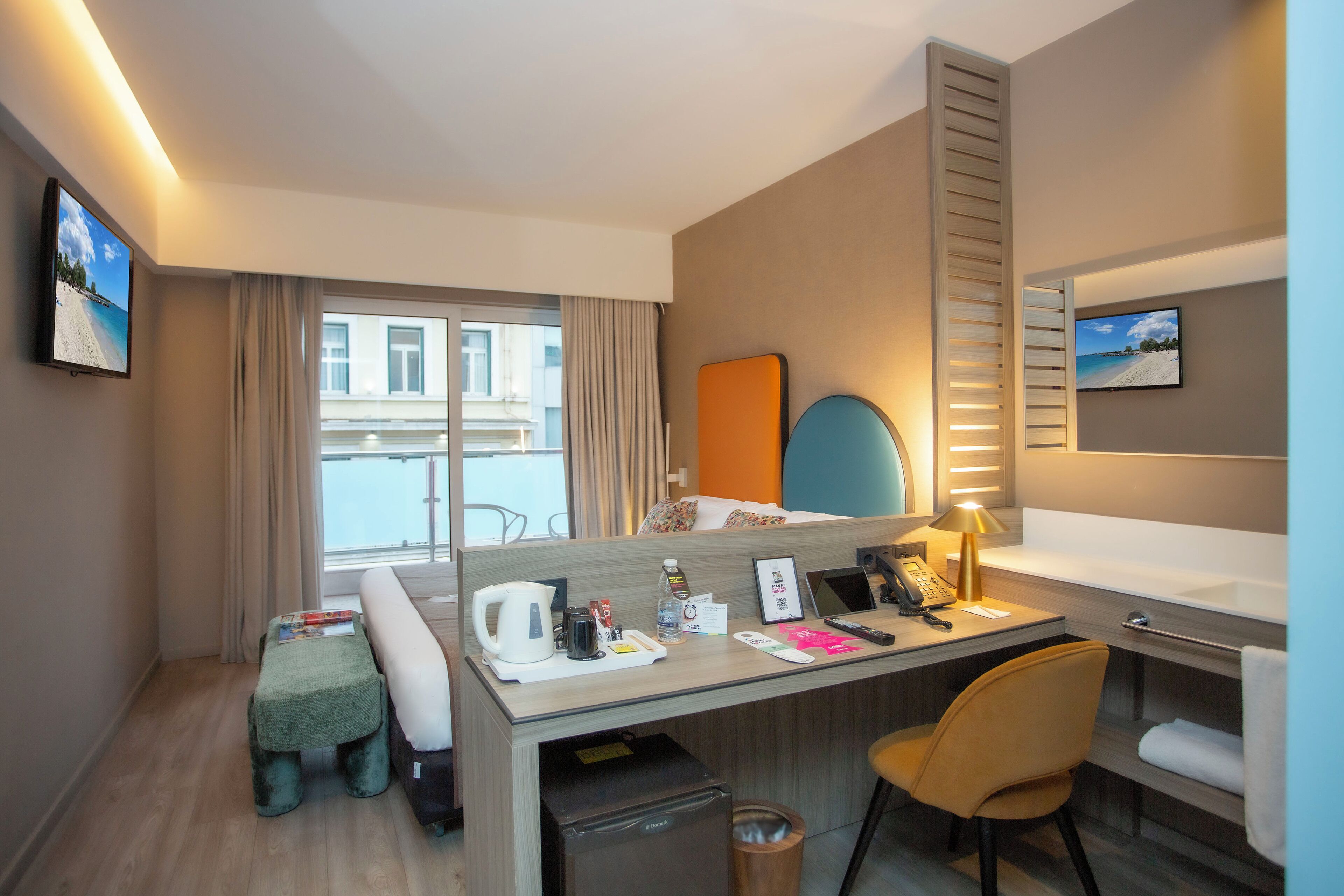 Een hotelkamer met een bureau, stoel en een bed. Er is een raam met gordijnen, een kleine tafel met een waterkoker en een fles, en een prullenbak.