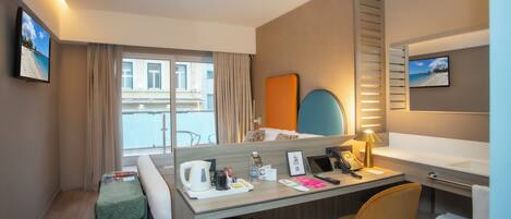 Een hotelkamer met een bureau, stoel en een bed. Er is een raam met gordijnen, een kleine tafel met een waterkoker en een fles, en een prullenbak.
