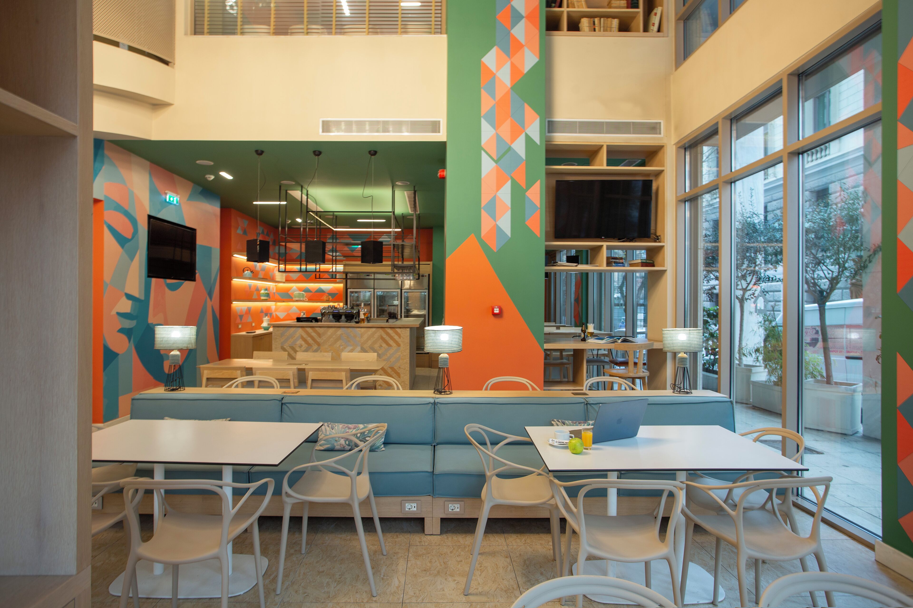 A modern dining area with a high ceiling, a central seating area with a blue cushioned bench, white tables, and chairs.