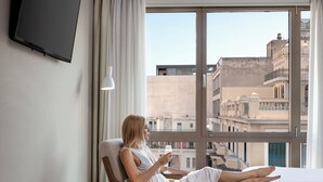 Une chambre d’hôtel avec une grande fenêtre, un lit, une chaise, une petite table avec une pomme et un verre, et un téléviseur à écran plat fixé au mur.