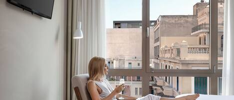 A hotel room with a large window, a bed, a chair, a small table with an apple and a glass, and a flat-screen TV mounted on the wall.