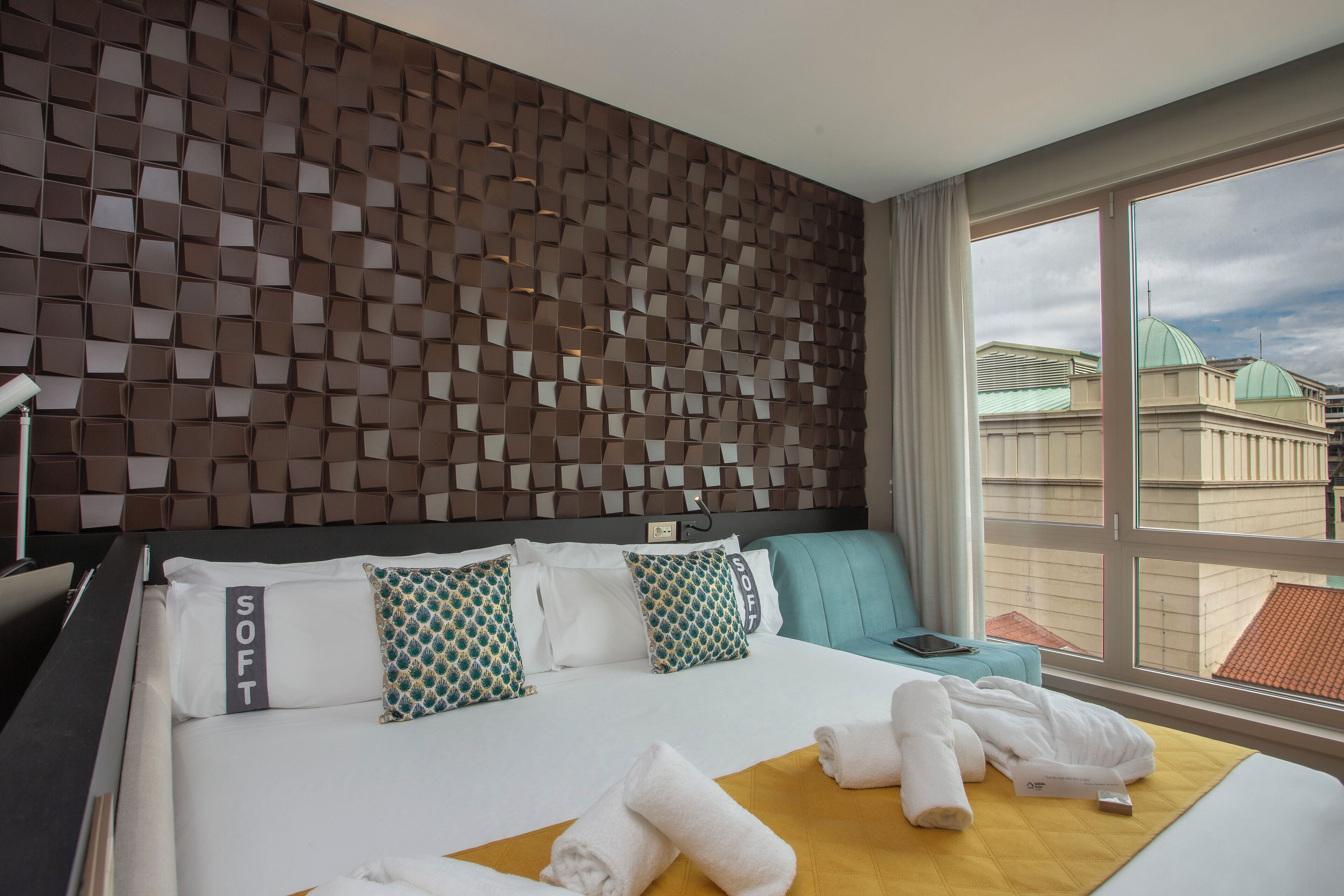 Quarto de hotel com design moderno, apresentando uma parede estampada, uma cama grande com várias almofadas, uma mesa de cabeceira e vista para prédios através de uma grande janela.