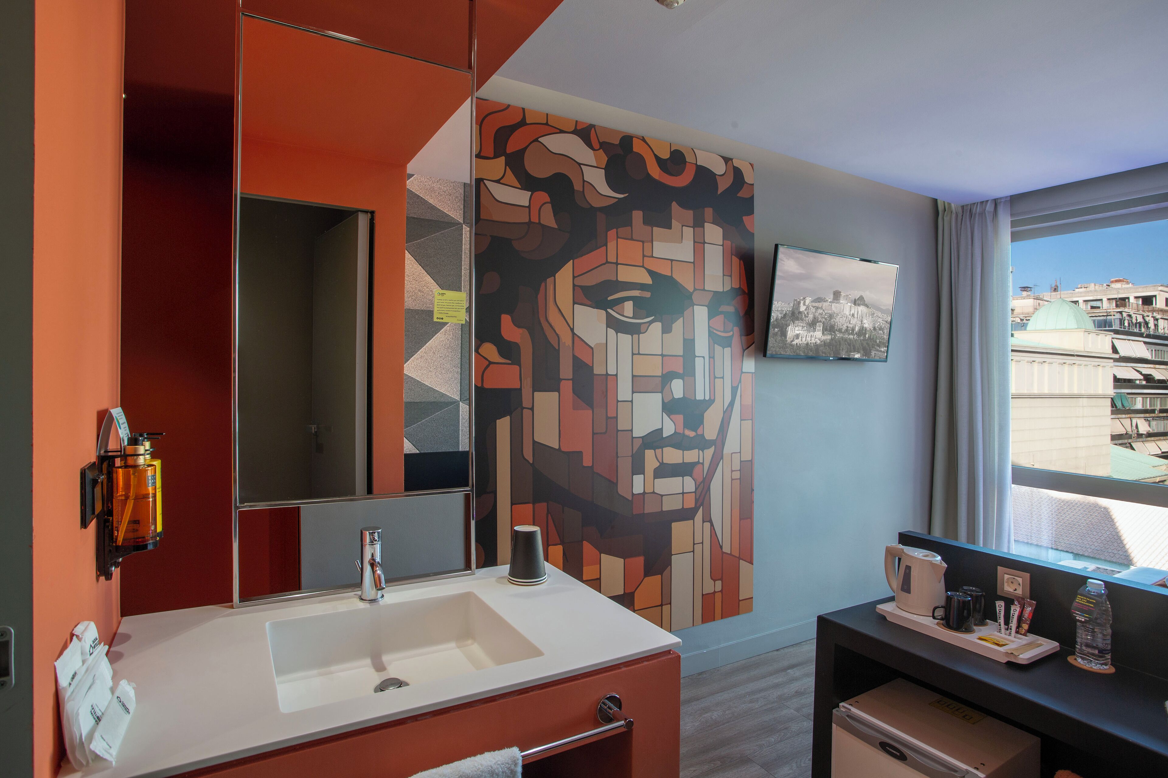 A modern bathroom with a large mirror, a sink, and a wall-mounted toilet. There is a window with a view of buildings and a cityscape.