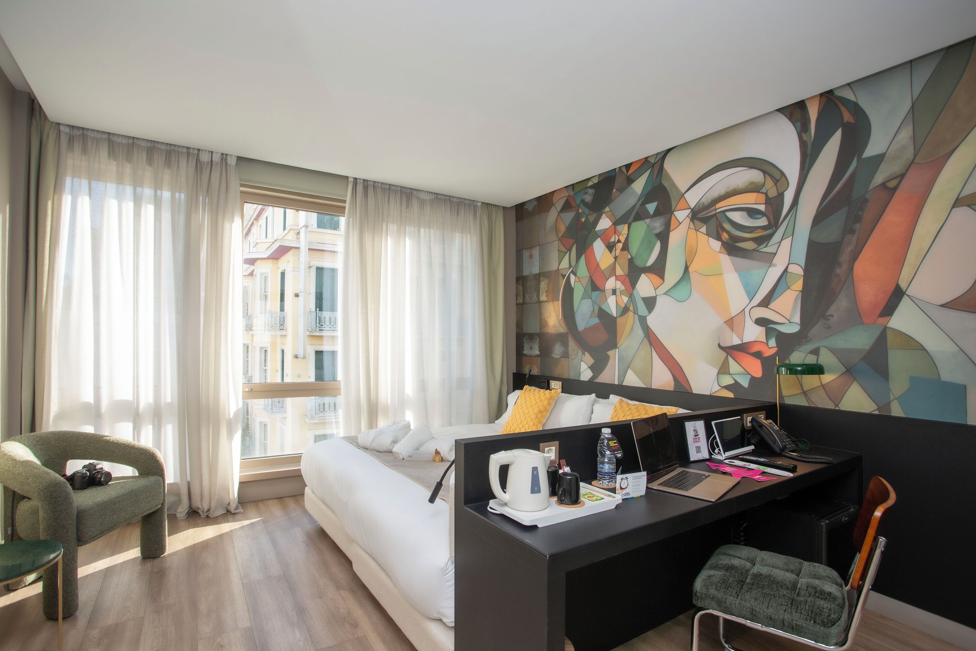 Un hotel moderno con una cama, un escritorio con una computadora portátil, una silla y un cuadro abstracto grande en la pared.