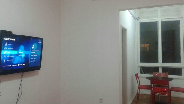 A room with a flat-screen TV mounted on the wall displaying a music streaming service interface. There is a window with white blinds, a red chair, and a small table. - Studio Maximo (Rio de Janeiro)