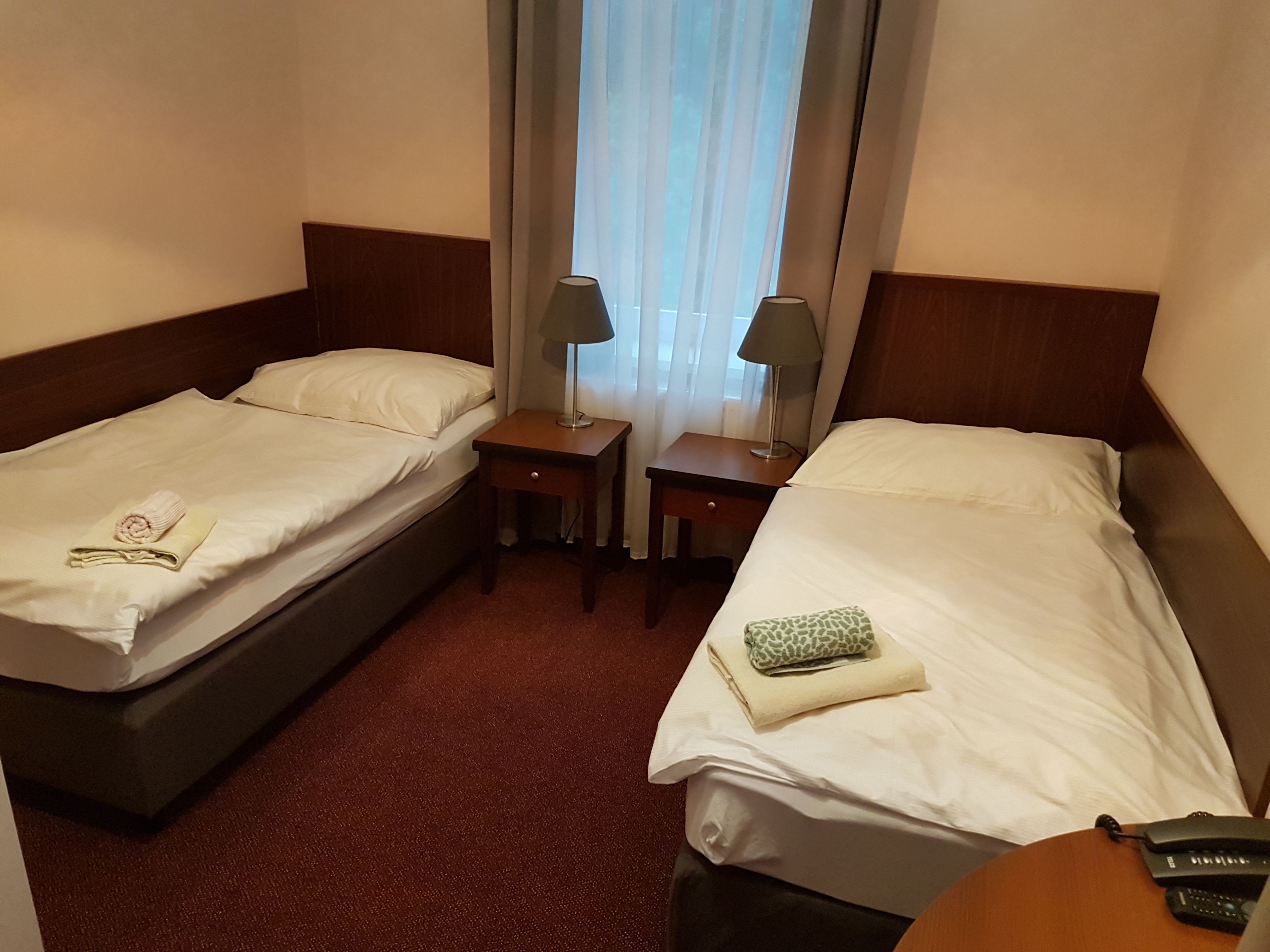 A hotel room with two single beds, each with a pillow and a folded towel. There are two bedside tables, one with a lamp and the other with a phone. The room has a wooden headboard and a window with sheer curtains.
