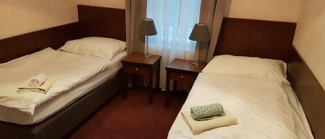A hotel room with two single beds, each with a pillow and a folded towel. There are two bedside tables, one with a lamp and the other with a phone. The room has a wooden headboard and a window with sheer curtains.