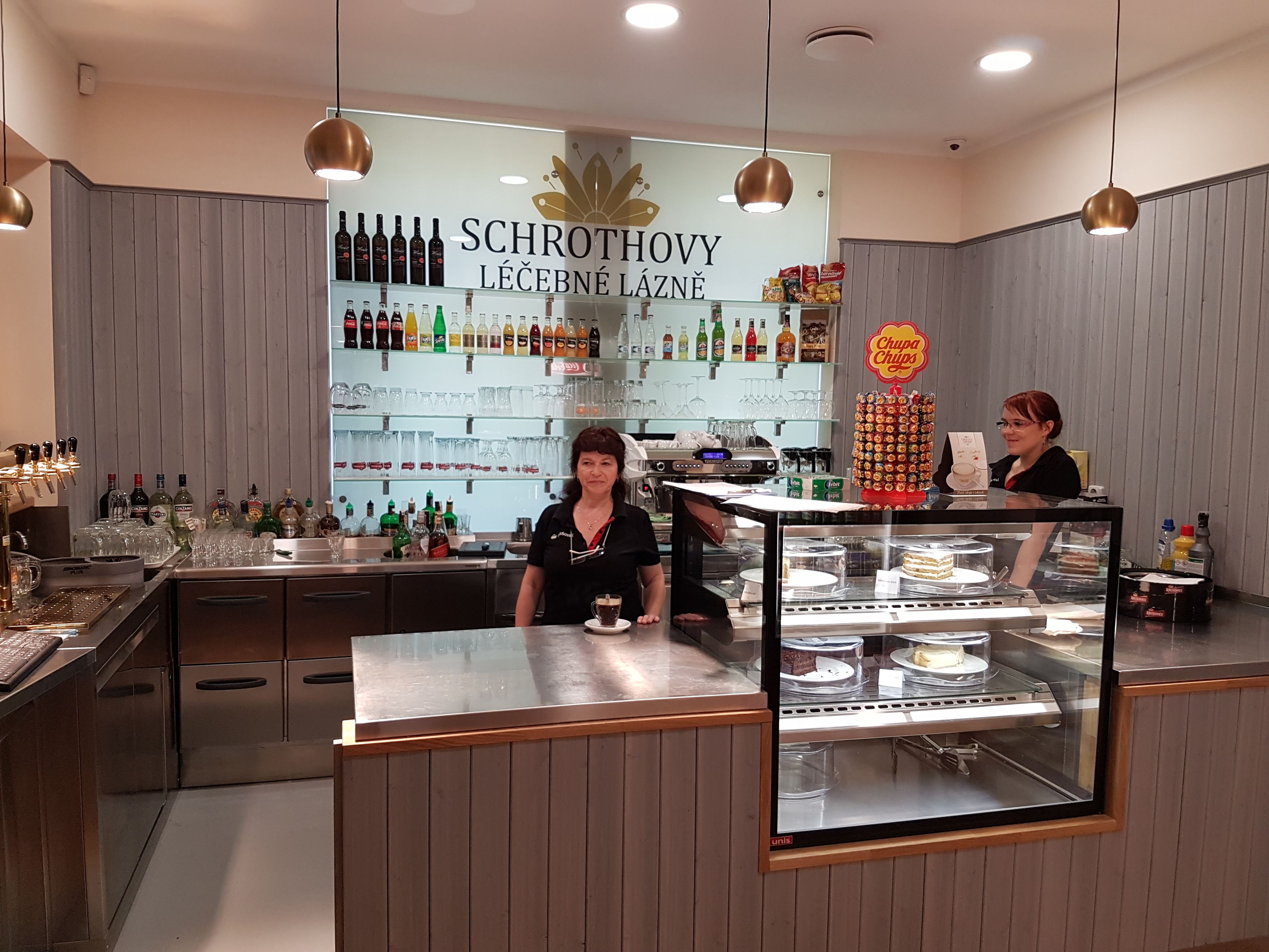 A bar area with shelves of liquor bottles, glasses, and a display case with desserts.