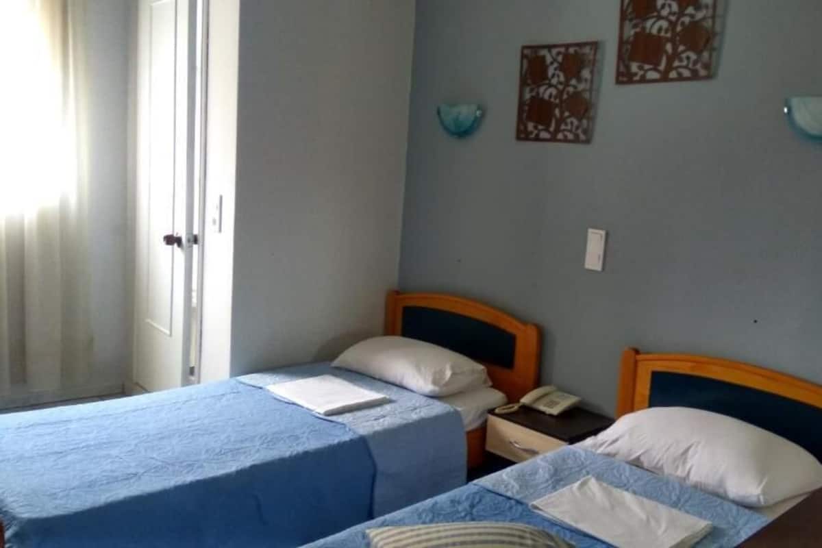 A room with two single beds, each with a blue bedspread and white pillows. There is a bedside table with a lamp and a phone. The walls are painted light blue, and there are two wall-mounted lamps.