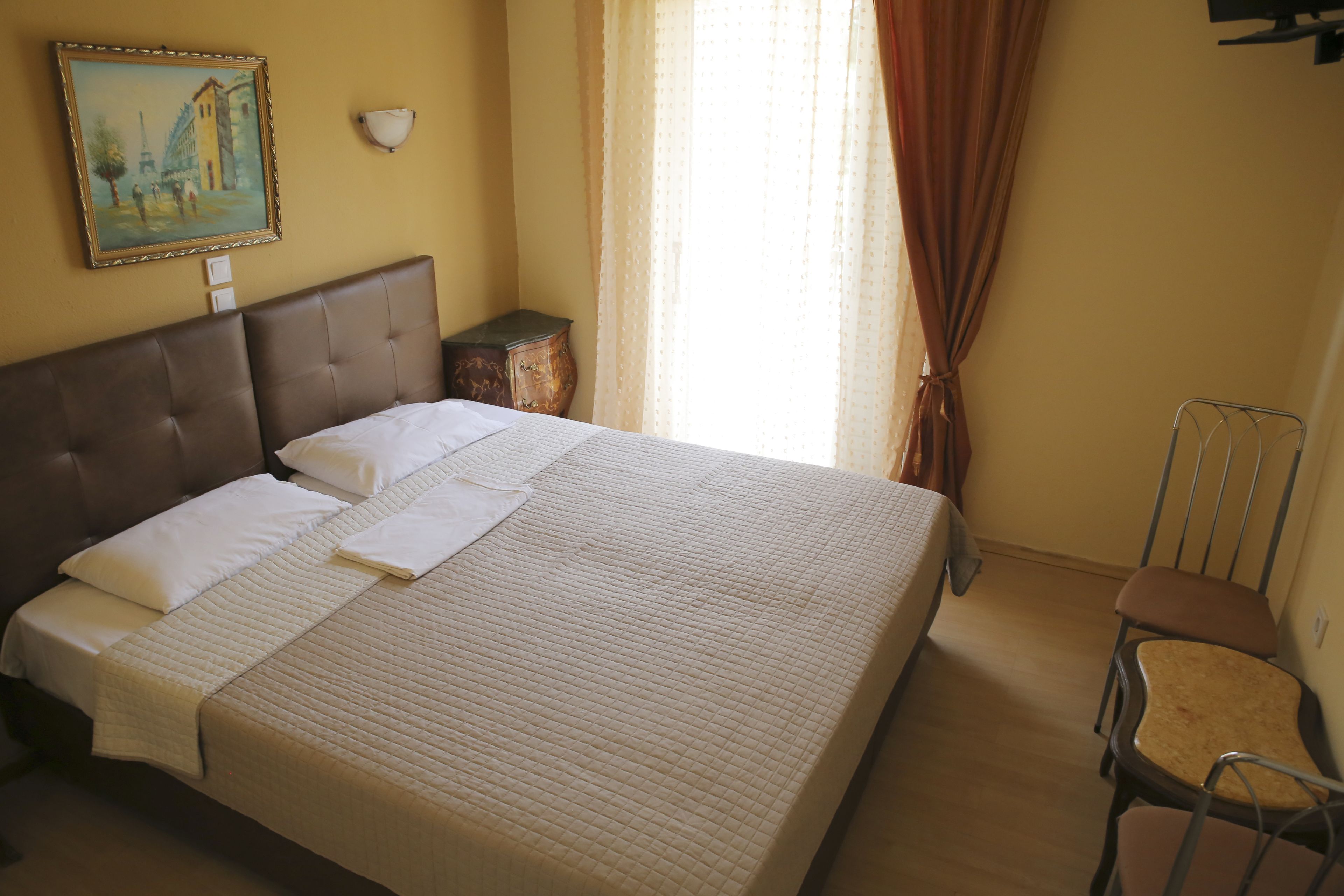 Uma cama bem arrumada com lençóis brancos, uma cabeceira, uma mesa de cabeceira com abajur e uma cadeira.
