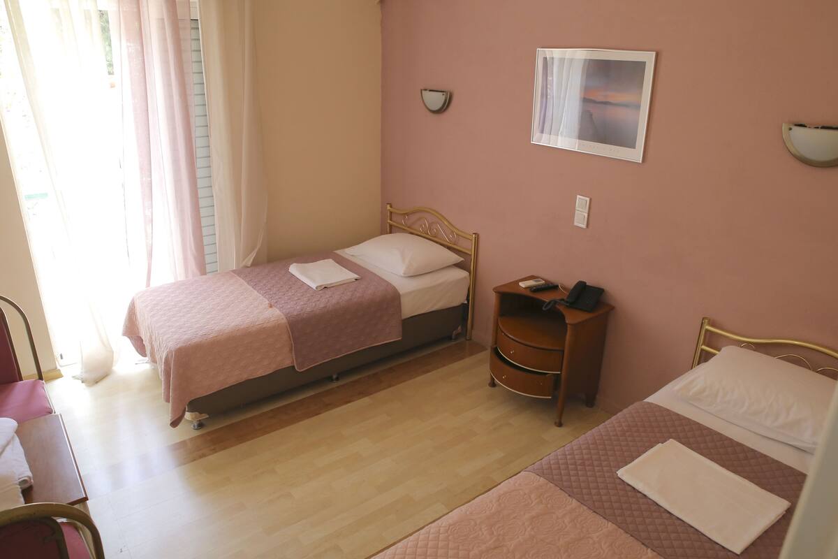 A room with two beds, a bedside table with a lamp, a wall-mounted light, and a window with curtains.