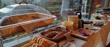 A buffet with various breads and pastries, a knife and fork on a plate, and a view of buildings in the background.