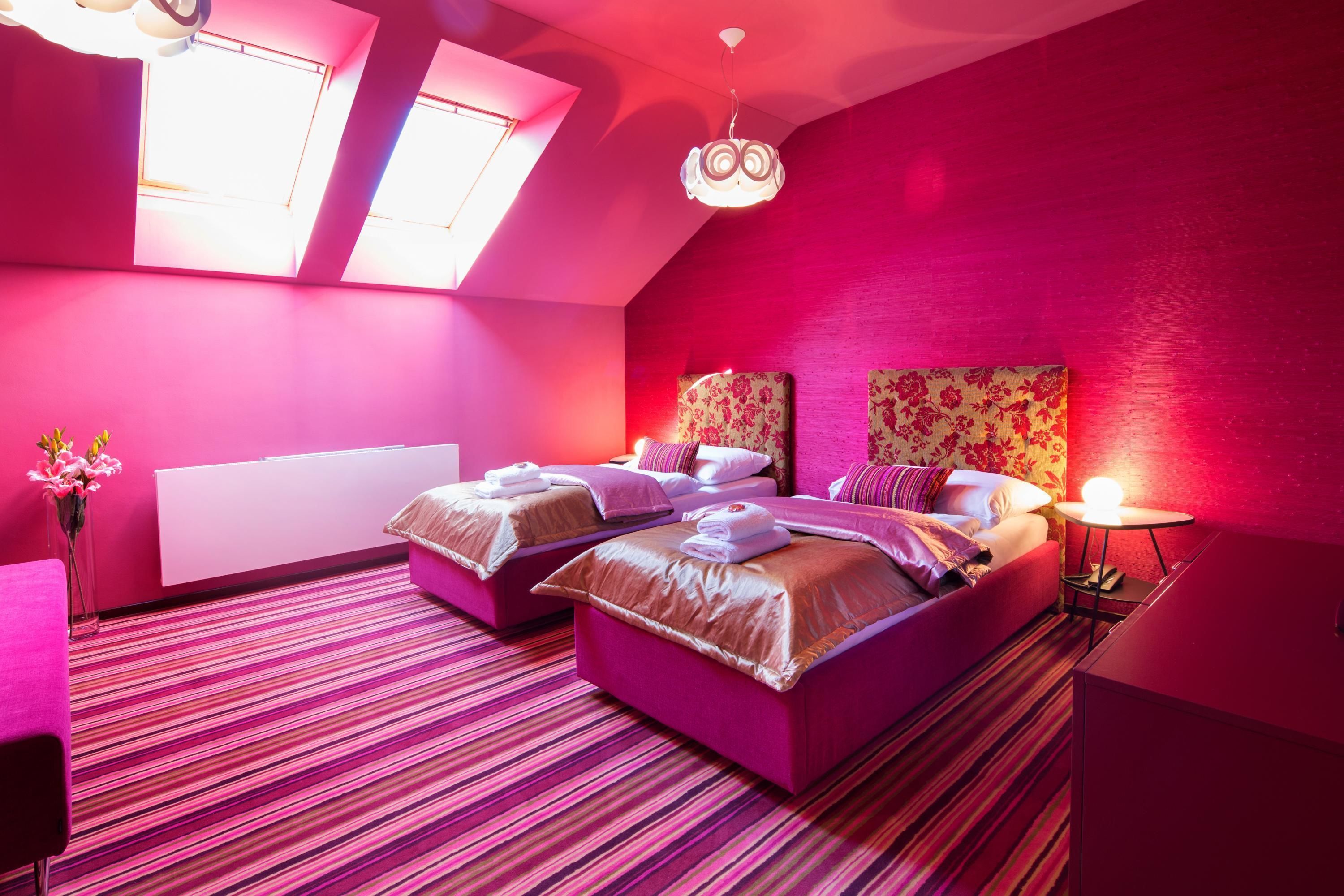 Una habitación con dos camas, una mesita pequeña y una mesita de noche. Hay dos ventanas con tragaluces, y las paredes están pintadas de rosa.
