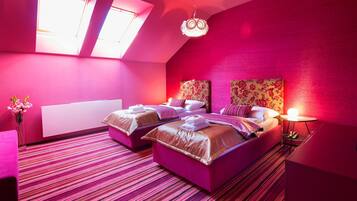 A room with two beds, a small table, and a nightstand. There are two windows with skylights, and the walls are painted pink.