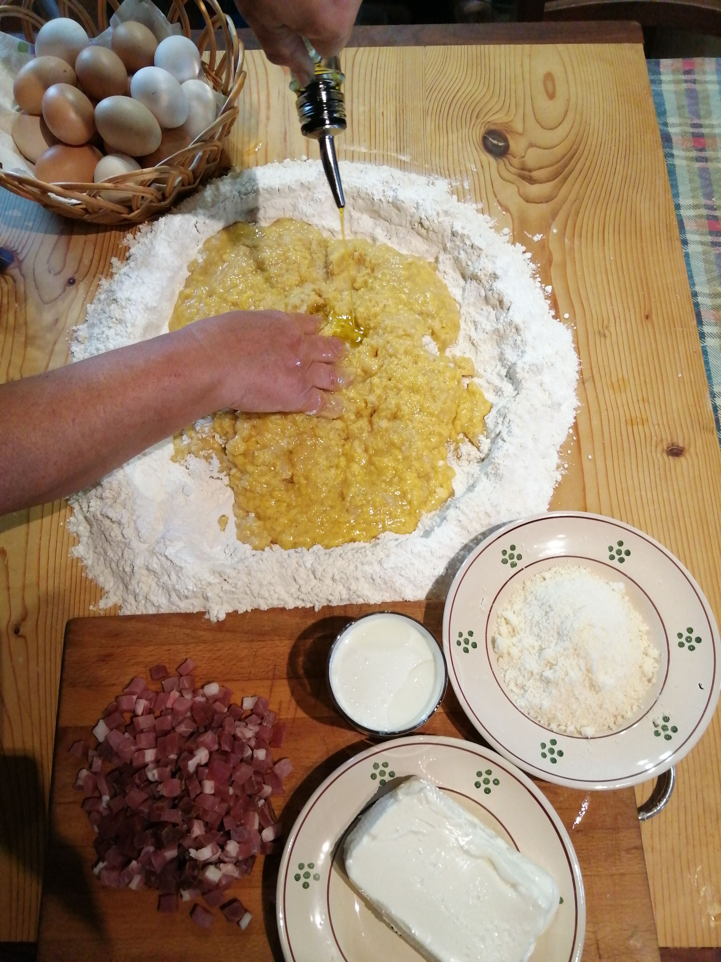 Una persona mezclando ingredientes sobre una mesa de madera, con un cuenco de huevos, un plato de jamón en cubitos y recipientes con harina y queso crema.