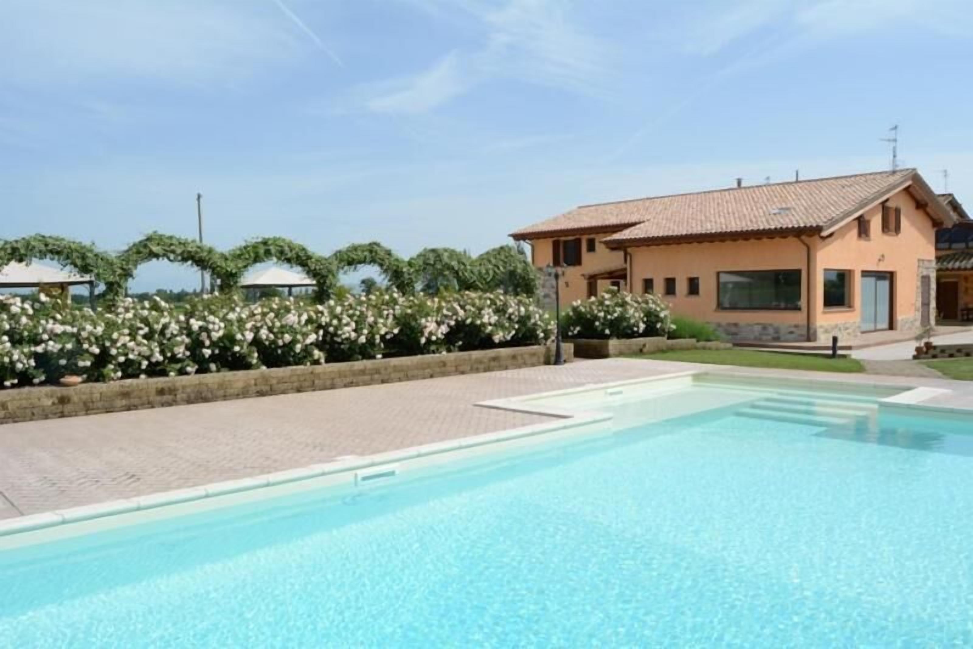 A swimming pool with a clear blue water surface, a building with a tiled roof, and a garden with white flowers and green foliage.