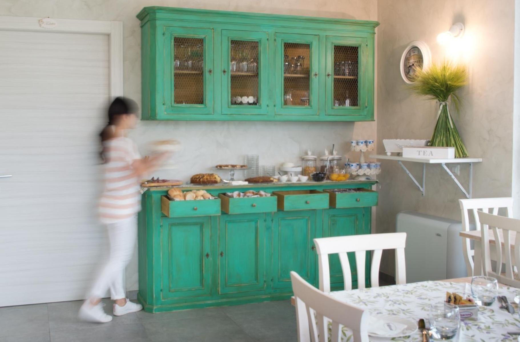 Una cocina con gabinetes color turquesa, una encimera blanca con diversos alimentos y una zona de comedor con sillas y una mesa blancos.