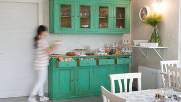 Una cocina con gabinetes color turquesa, una encimera blanca con diversos alimentos y una zona de comedor con sillas y una mesa blancos.