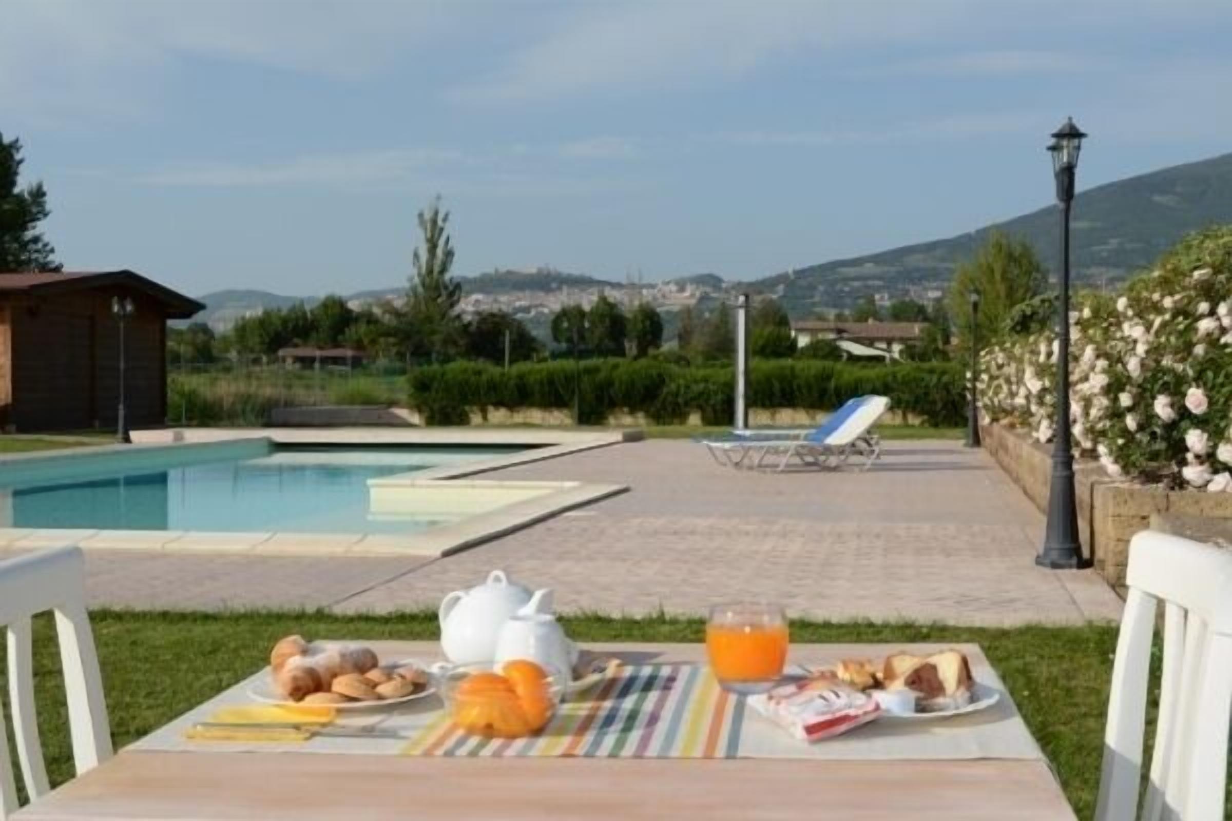 A breakfast setup with croissants, orange juice, and a teapot on a table with a view of a swimming pool and a mountainous landscape.