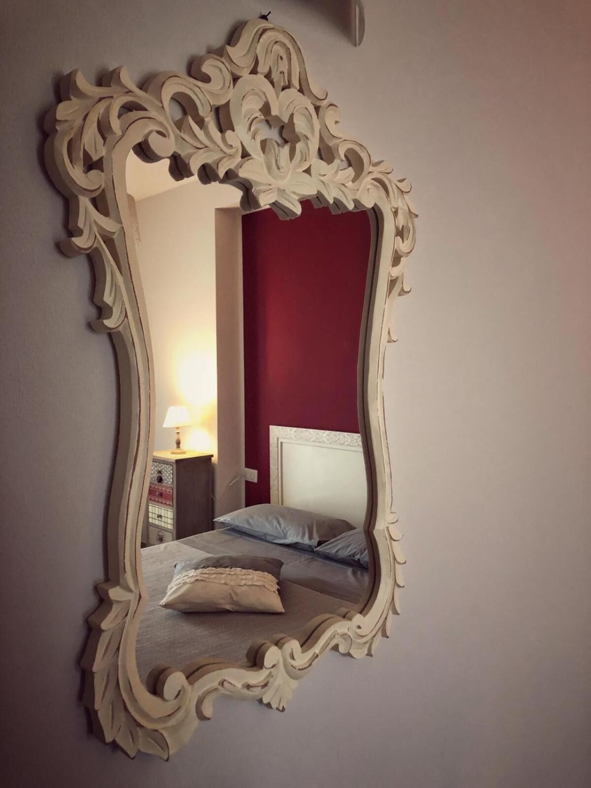 Ein Spiegel mit aufwendiger Rahmen, der einen Raum mit einem Bett, Kissen und einem Nachttisch mit Lampe reflektiert.